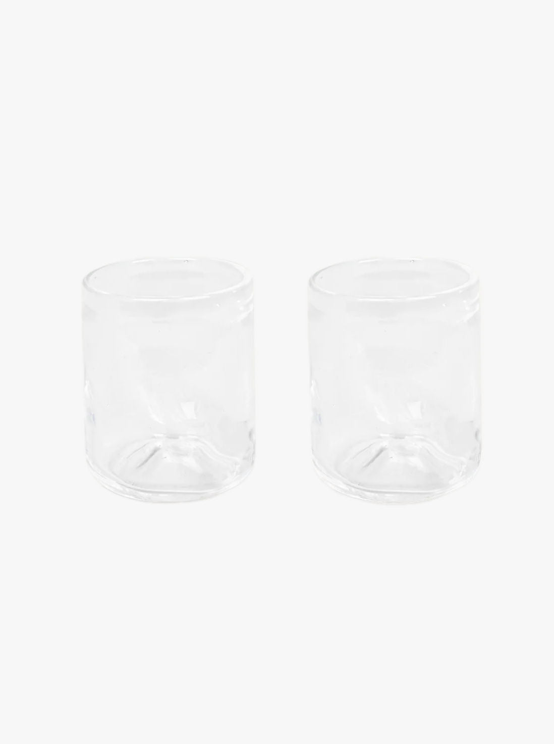 Two clear, cylindrical 0405 Glass Set of Two | Clear | Small candle holders featuring a handblown design by FRAMA are placed side by side on a plain white background. They are empty and have smooth, even surfaces.