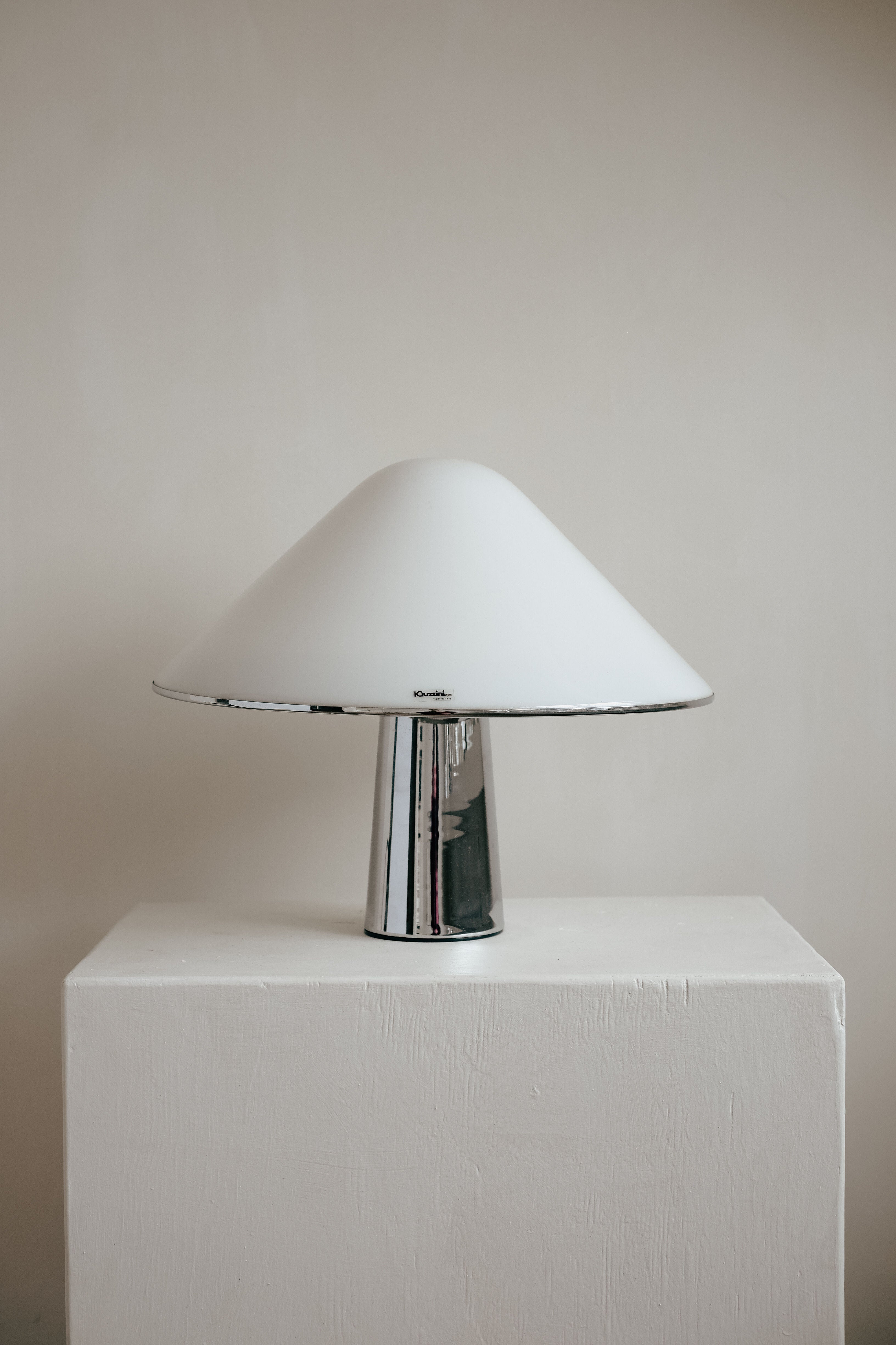 The Harvey Guzzini Elpis Table Lamp by septembre studios, featuring a sleek metallic base and smooth white dome-shaped shade, is reminiscent of Luigi Massoni's design. It rests on a plain white pedestal against a neutral background.