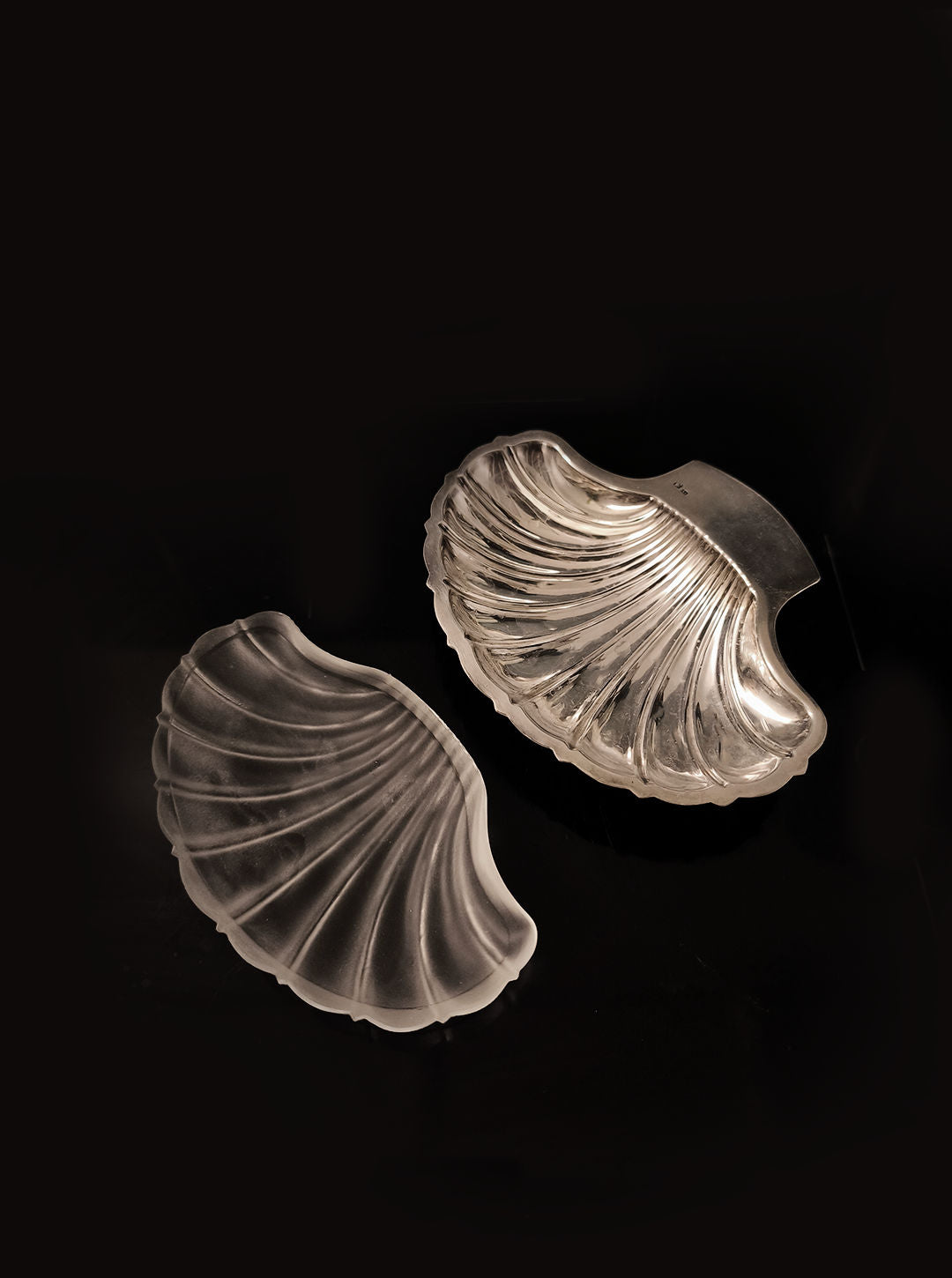 Displayed against a black background, two transparent textured glass objects by Les Objoies resemble seashell halves. Their intricate ridges and glossy finish evoke elegance, making them the perfect Shell Butter Dish to enhance any table setting.