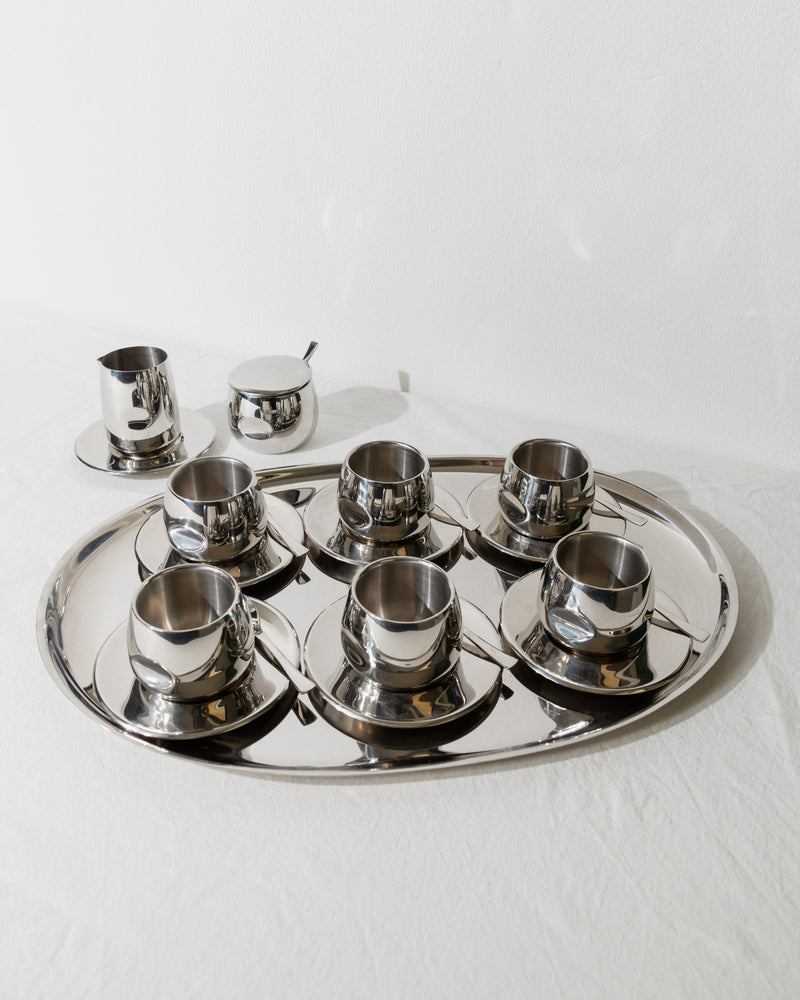 A Bottega Jacobs masterpiece, the Vintage Coffee Set - Italy 70s, crafted with polished steel, gracefully adorns the tray. It includes six stylish cups with saucers, a small creamer, and a sugar bowl with a spoon. This elegant set rests on a white tablecloth against a plain background.