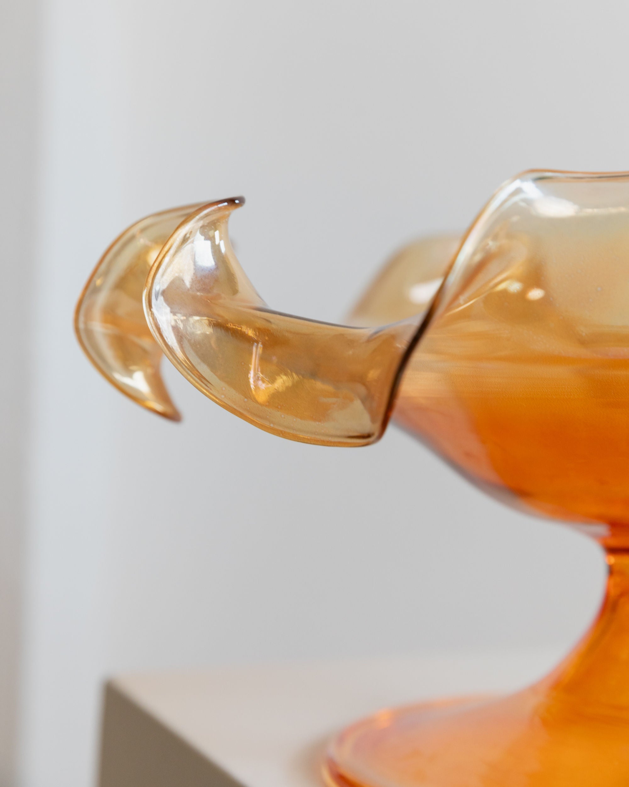 A close-up of the Murano Glass Bowl by Parise Vetro 70s from Bottega Jacobs reveals a decorative piece reminiscent of vintage designs, featuring swirling, translucent amber curves and fluid shapes. Its smooth, glossy surface boasts an orange iridescence with delicate edges emerging from a central base against a neutral background.