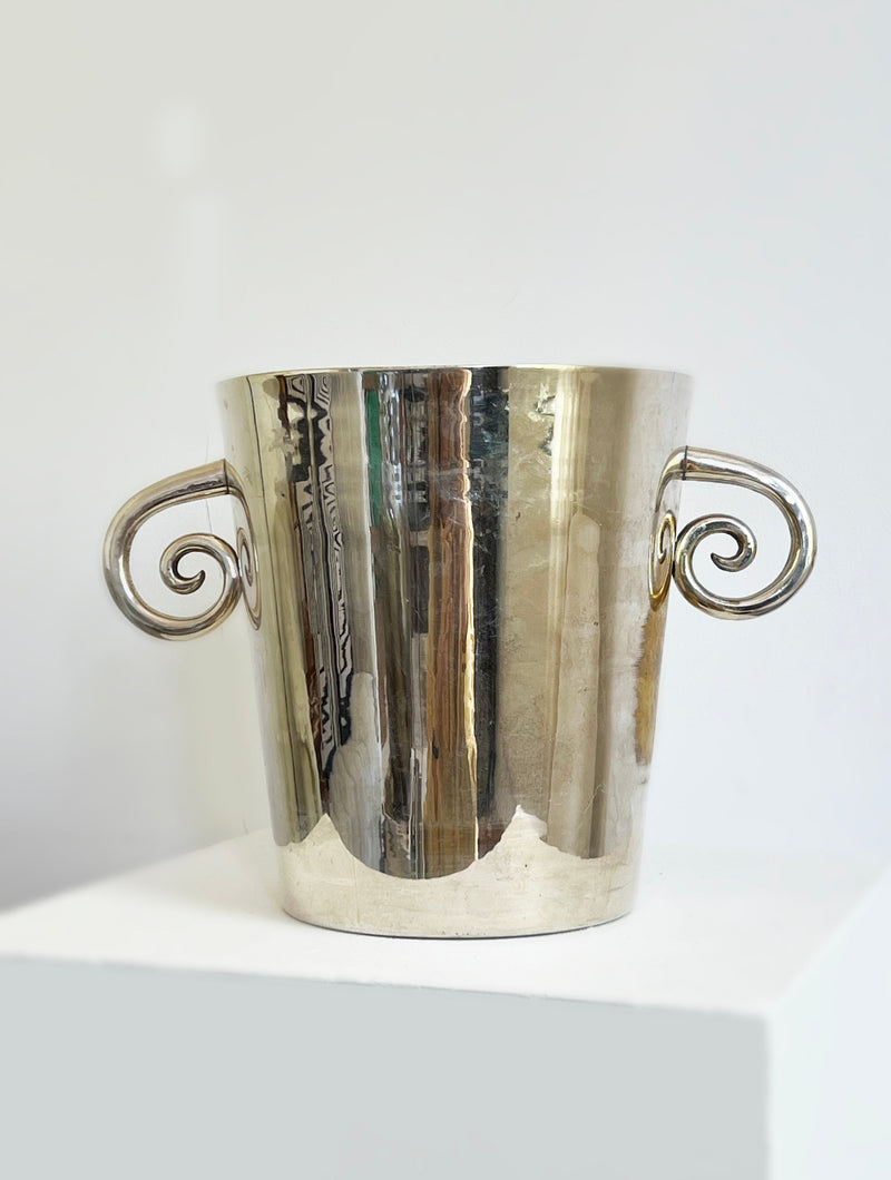 The Porter Studio Spiral Handle Ice Bucket, featuring shiny silver finish and ornate curled handles, is displayed on a white pedestal. Its reflective surface subtly mirrors the plain background.