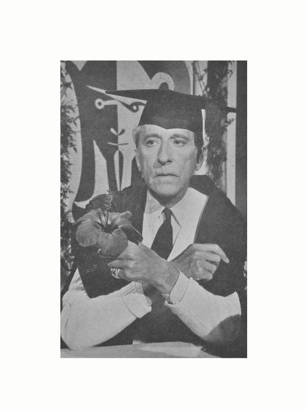 A black-and-white photo of an older man wearing a graduation cap and gown. He is sitting with his arms resting on a table, holding a plant with large leaves and a flower. Behind him is the Spécial Cocteau Vintage Book by Maison Plage, evoking the charm of a vintage book. The man appears to be deep in thought.