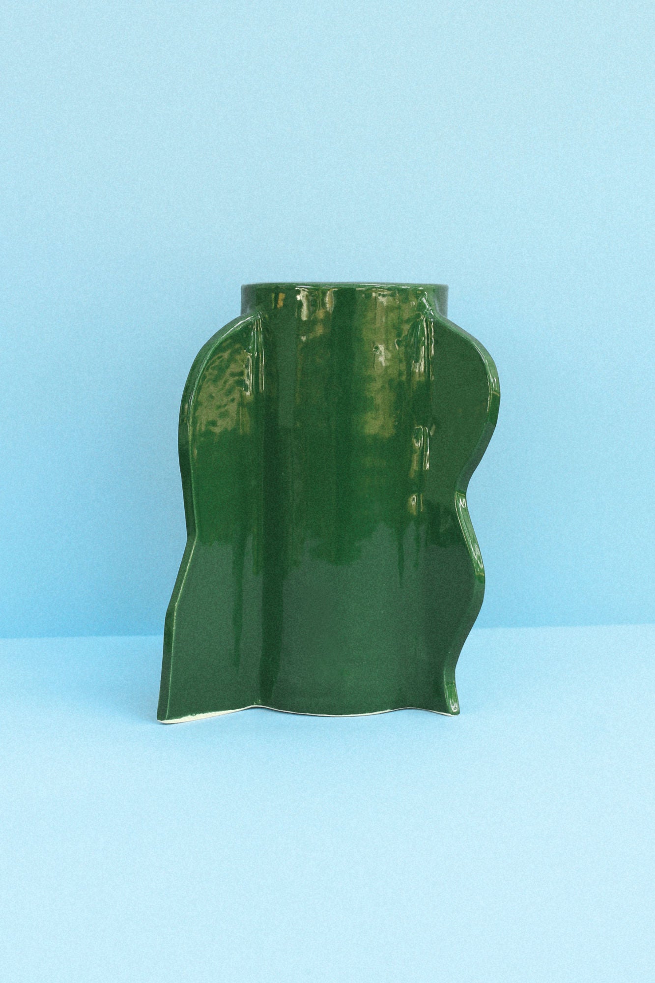 A green, irregularly shaped ceramic vase with a glossy finish set against a light blue background. The artisanal hand-painted *Theatre Vase 04 (Bosque)* from Casa Veronica has an abstract, wavy form, creating a unique and artistic appearance.