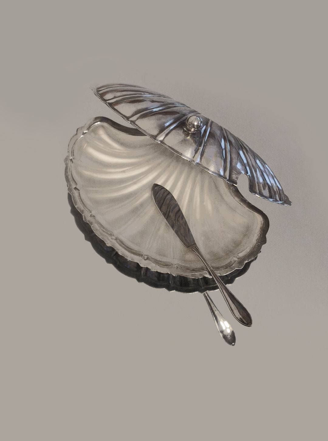 The Les Objoies Shell Butter Dish is silver-plated with a hinged seashell design, revealing a matching butter knife inside. Its glossy, reflective surface and intricately designed knife handle enhance elegant dining settings.