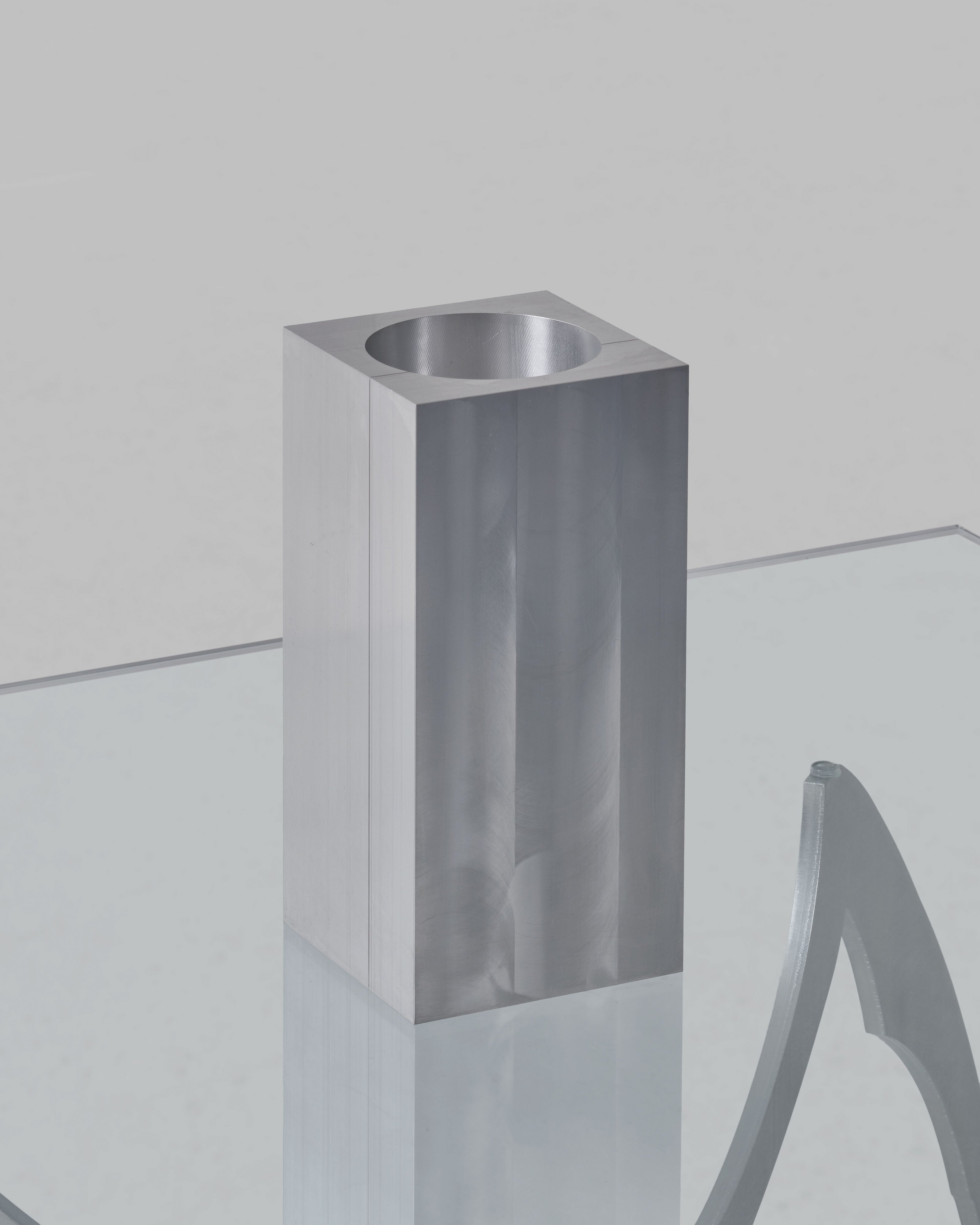 A minimalist, rectangular ALU Vase from 304.cage with a cylindrical opening sits on a transparent glass surface. The vase exhibits a sleek and polished aluminium finish, reflecting light and casting subtle shadows, while an abstract metallic sculpture is partially visible beside it.