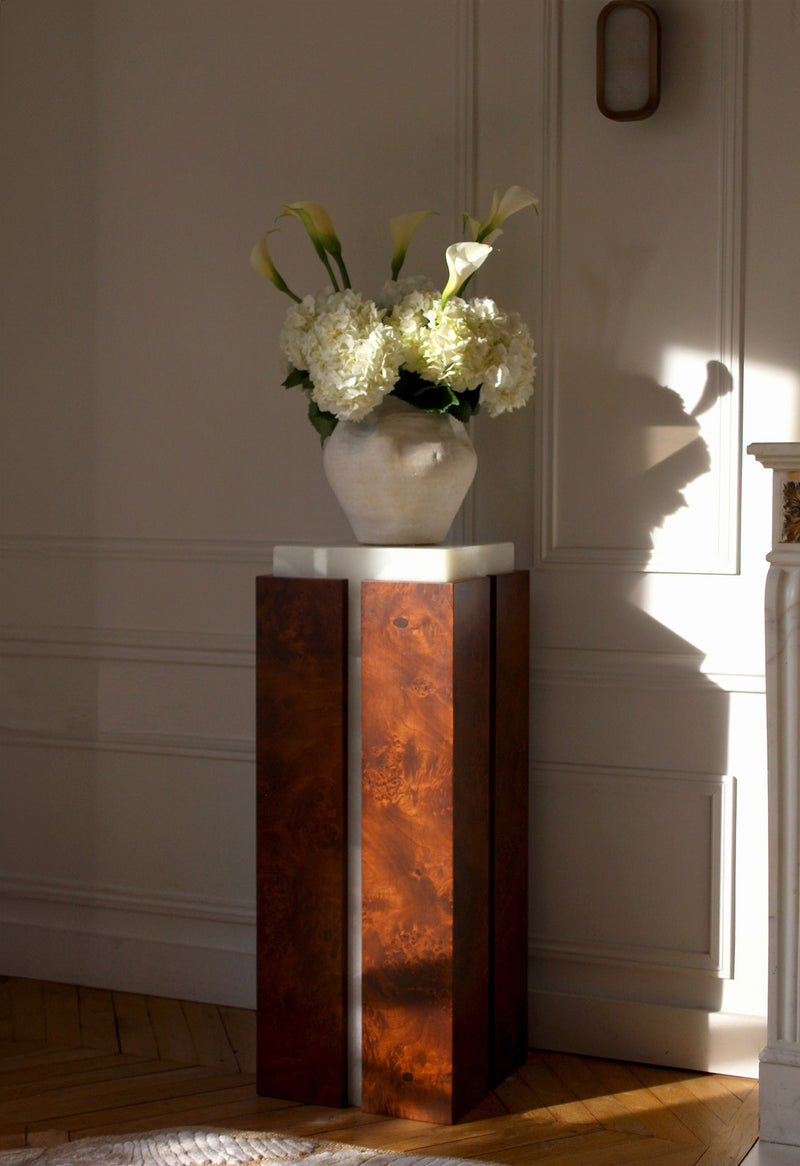 A white ceramic vase with hydrangeas and calla lilies sits on the Marbera 'Como' Plinth in white onyx and brown wood, placed in a sunlit room with white paneled walls and parquet flooring.