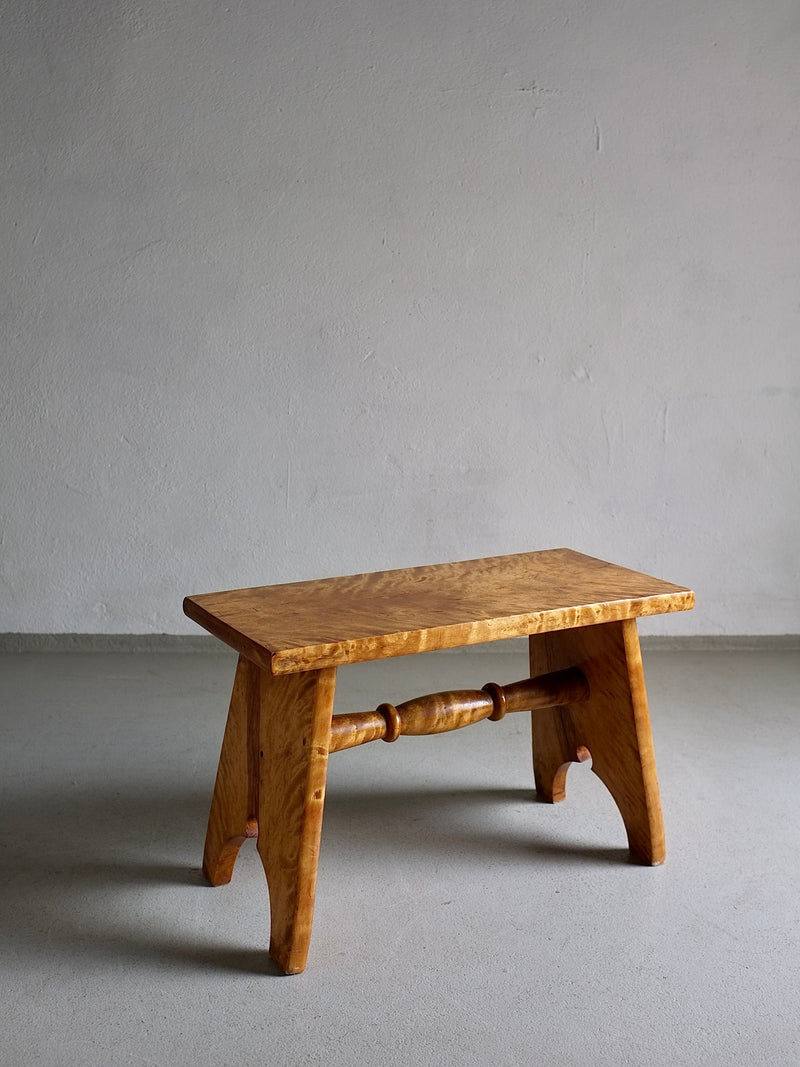 A small, handcrafted wooden bench stands against a plain, gray backdrop. The Rustic Solid Karelian Birch Stool | Latvia | 1950s from Veter Vintage, resembling a carved low stool, features a simple design with a flat top and two angled legs connected by a round spindle. Made from solid Karelian birch wood in vintage condition, its light, natural finish highlights the grain.