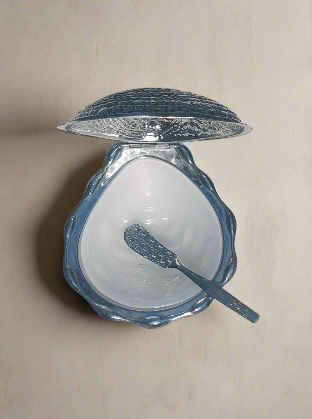 A silver caviar spoon rests inside an open Shell Butter Dish by Les Objoies, featuring a white interior and clam-shaped vintage design, placed on a marbled surface. The container's textured lid is positioned behind it, casting a shadow.