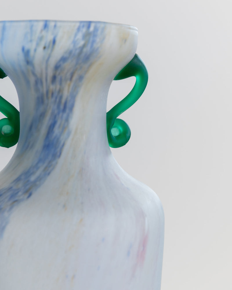 A close-up of the Murano Glass Vase 70s by Bottega Jacobs showcases a marbled blue and white pattern with vintage charm, enhanced by green decorative handles. The background is light and neutral.