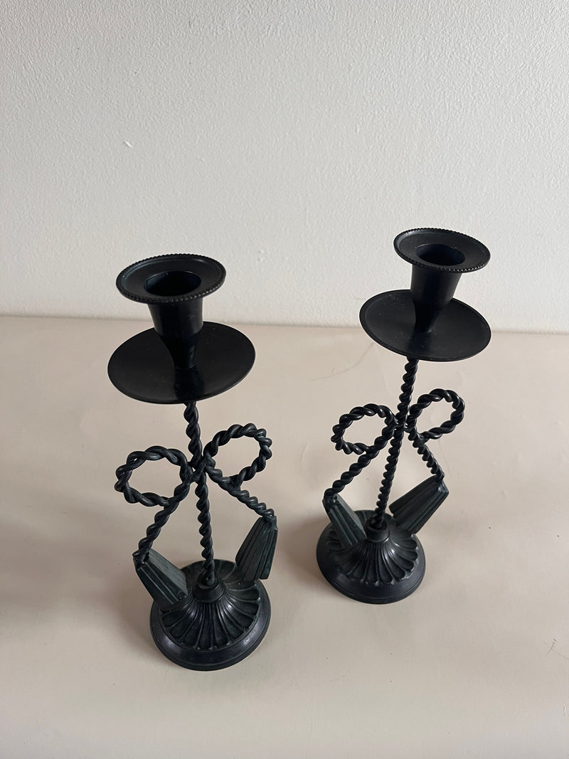 The Vintage Bow and Tassle Candleholders by Maud Vaughan, with twisted stems and intricate looped designs, grace a light surface. Elegant bow and tassel details enhance their charm against the plain white background.
