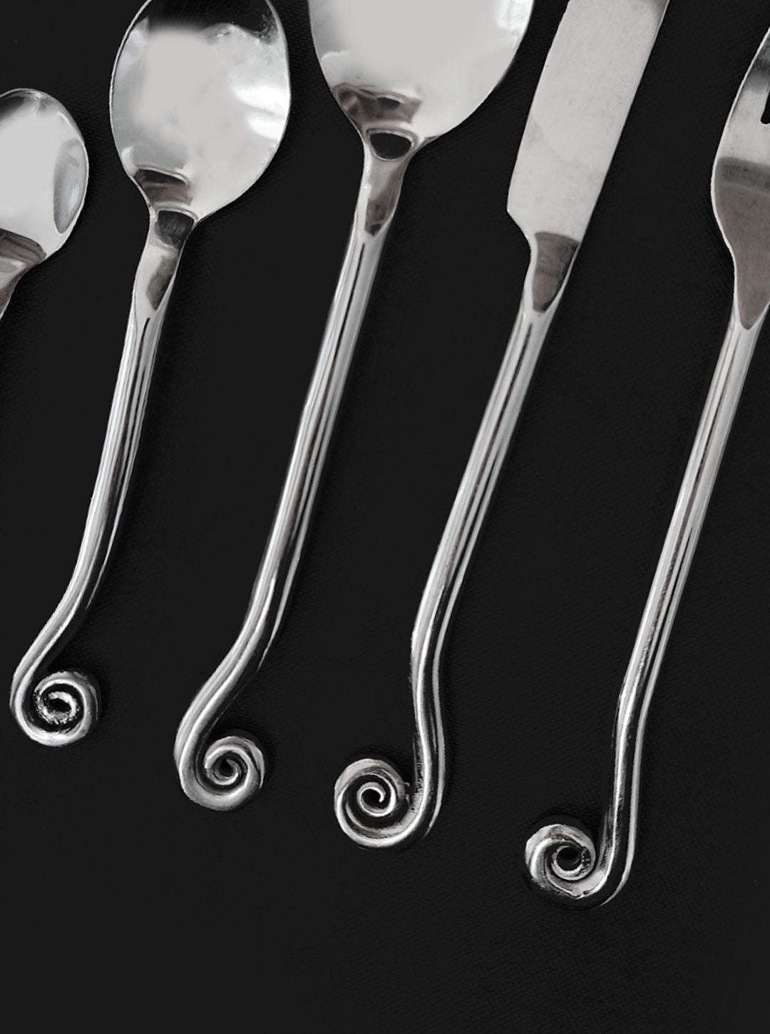 The Les Objoies Set of 5 Swirling Cutlery showcases a contemporary silver spoon, knife, and fork with ornate spiral designs at the handle ends, elegantly arranged in a row against a dark background.