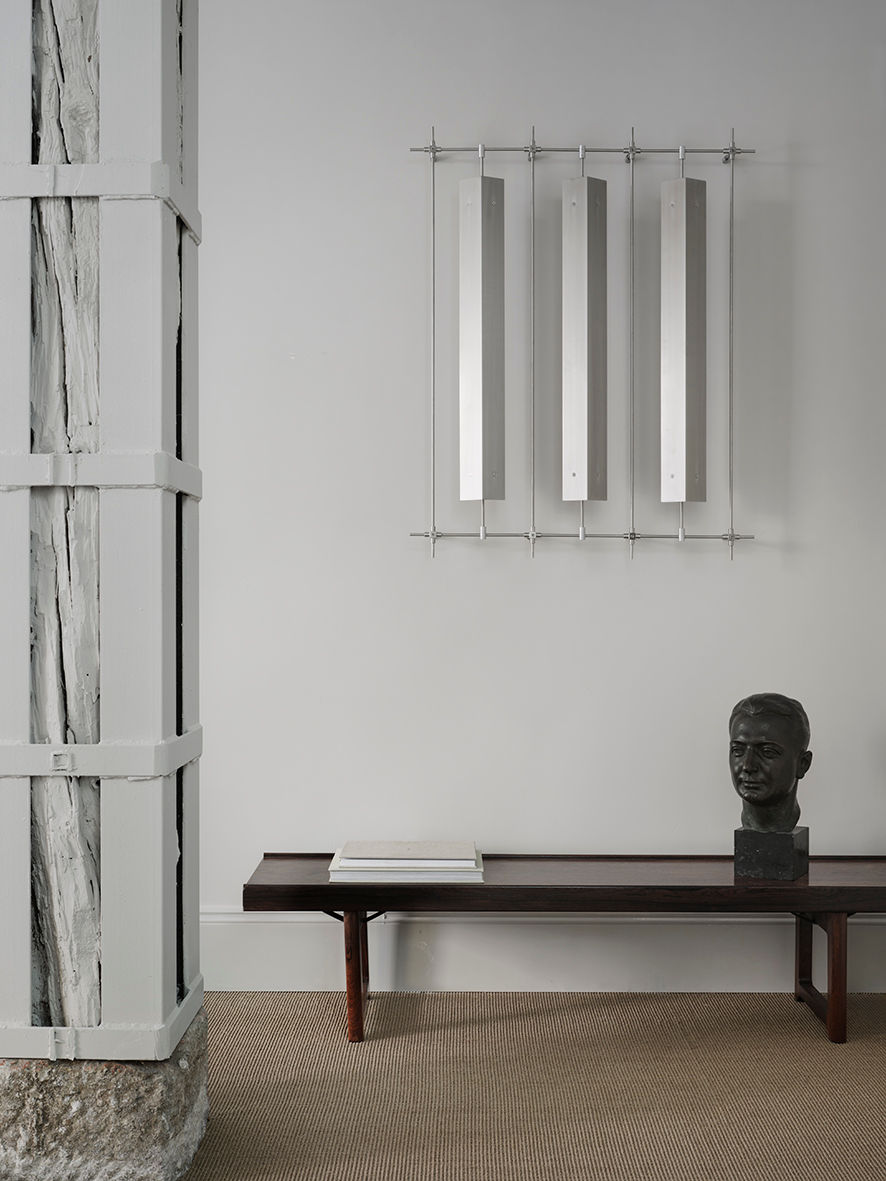 A minimalist room features the TESTA VIRILE modern bronze sculpture from FEBRERO SHOP and a low wooden bench. Three sleek, vertical metal panels hang on the wall. The floor is carpeted, and a rustic beam is visible on the left.