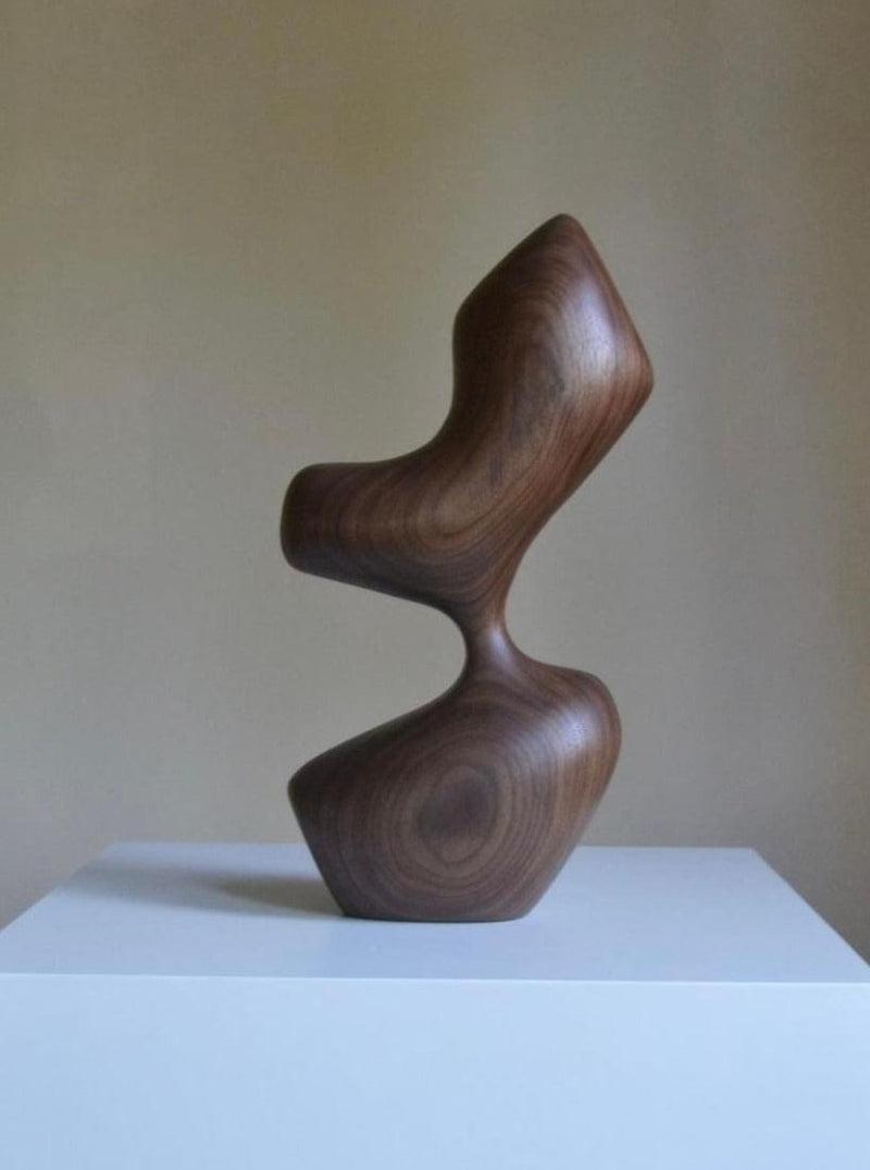 An Arbol wooden sculpture by Chandler McLellan, featuring a smooth, twisted design resembling interconnected hourglass shapes, displayed on a white pedestal against a beige background.