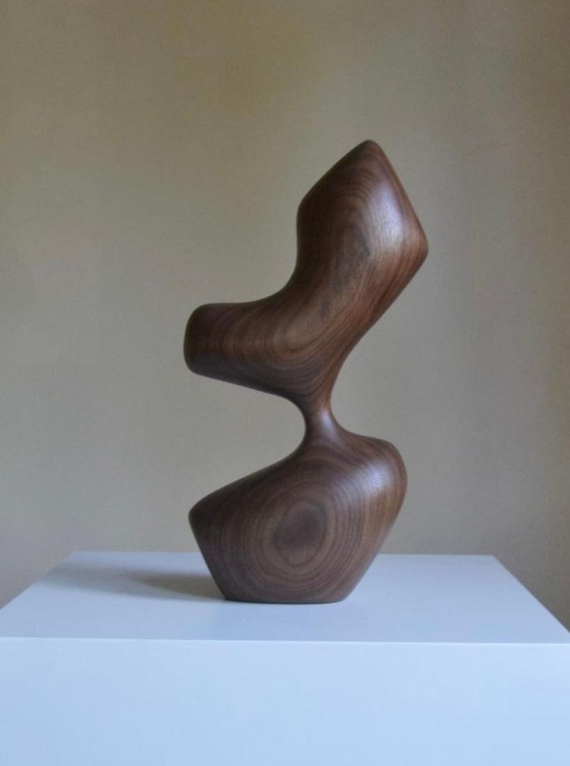 An Arbol wooden sculpture by Chandler McLellan, featuring a smooth, twisted design resembling interconnected hourglass shapes, displayed on a white pedestal against a beige background.