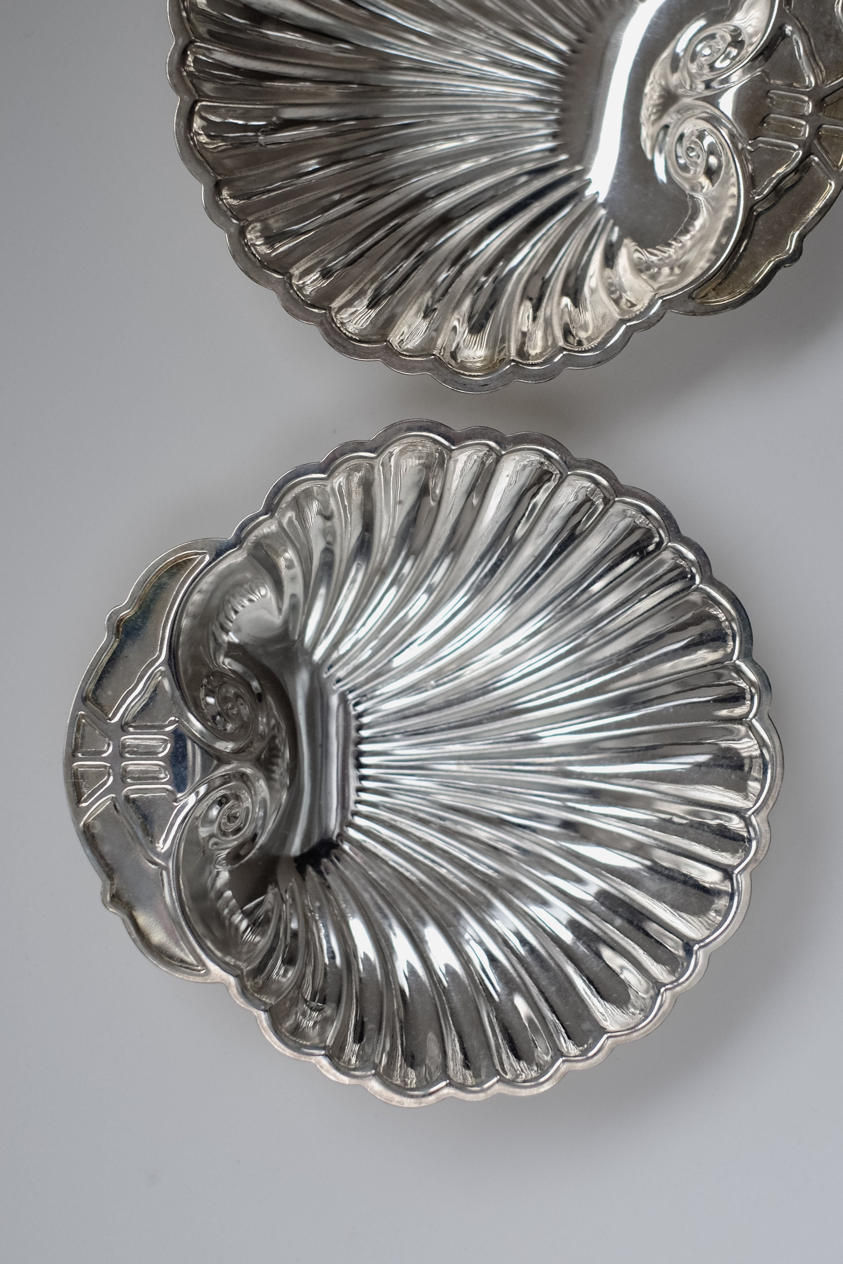 The Set of 2 Silver-plated Shell Trays by septembre studios, featuring ornate, vintage-inspired designs and intricate edge details, are artistically arranged with one tray partially overlapping the other on a plain white background, conjuring a refined dining ambiance.