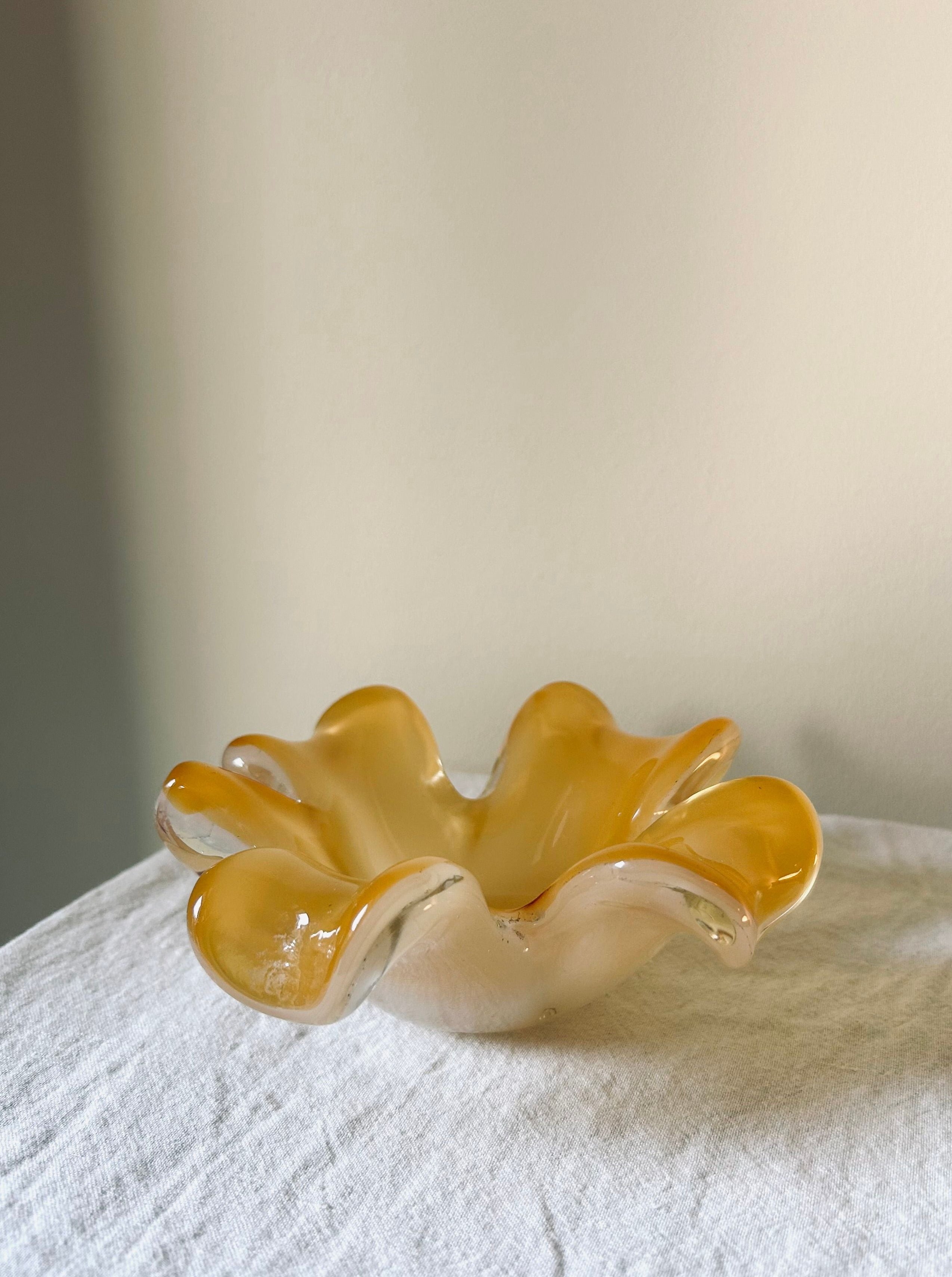 A dynamic Murano Glass Dish by Flock Studio, featuring wavy edges and a golden yellow hue, sits on a white surface. Its curvy design makes it an exquisite objet d'art against the neutral background.