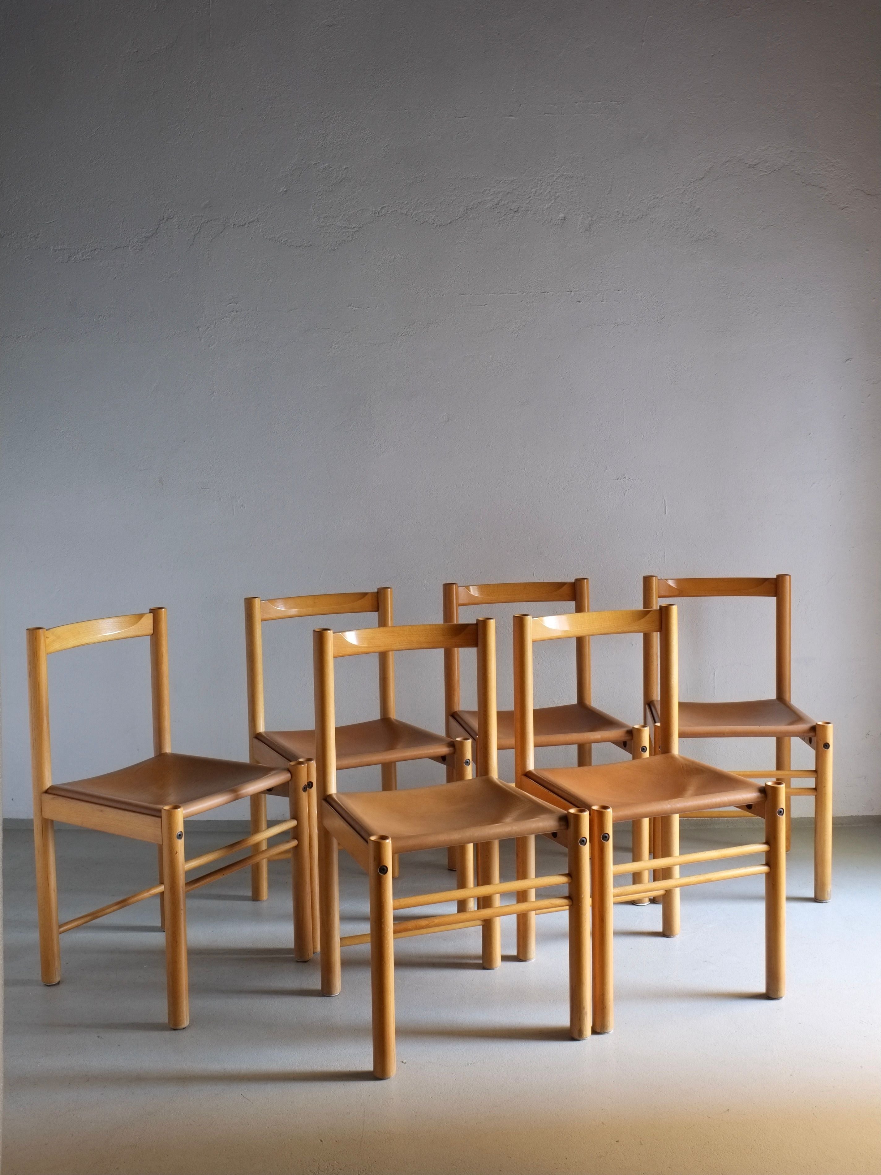 In a softly lit room with a plain, light-colored wall and floor, six Ibisco dining chairs from the 1960s by Veter Vintage are elegantly arranged. Crafted from beech wood with cognac leather seats, these vintage pieces feature distinctive rectangular backs.