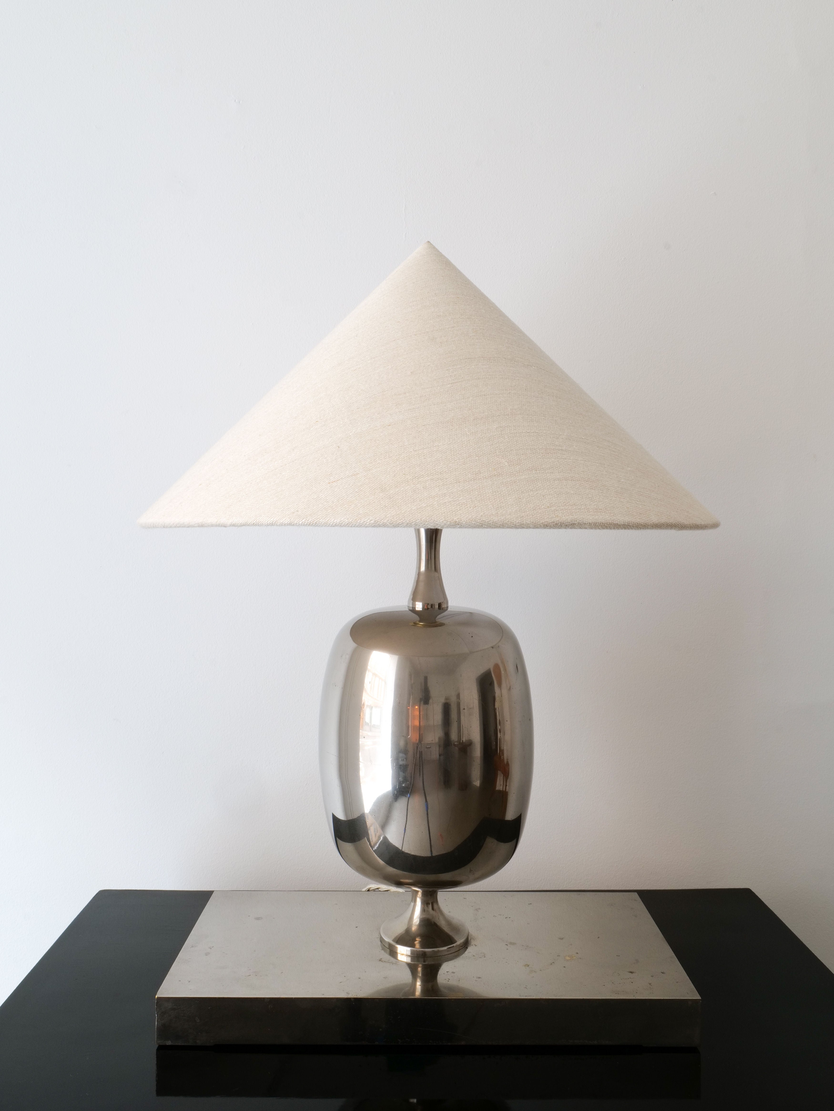 The Barbier Chrome Table Lamp by Collection apart features a reflective silver base that recalls Philippe Barbier's iconic chrome over brass designs, paired with a wide cream-colored conical shade. It's beautifully showcased on a glossy black surface against a plain white wall.