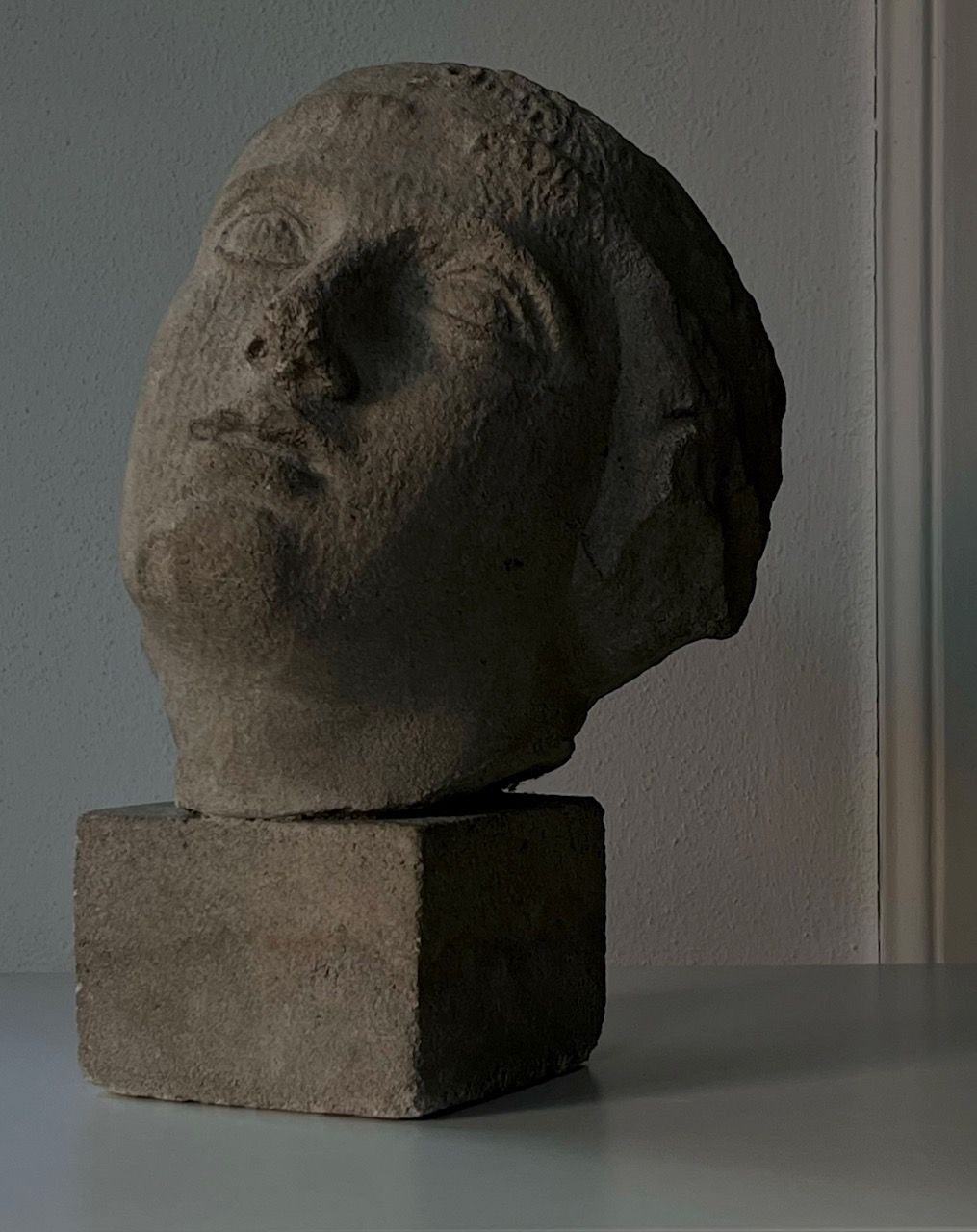 The Decorative Bust by Médecine features a contemplative face slightly tilted atop a square stone base, crafted from sandstone composite, and stands out beautifully against a simple, neutral backdrop.