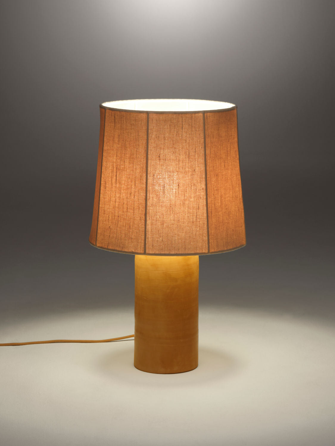 A warm beige-colored table lamp with a terracotta base and a pure linen lampshade is illuminated against a plain gray background. The Sonora Linen Table Lamp by Nassi Lamps features a design that is simple and modern, evoking the charm of Puglia, with the power cord visible extending from the base.