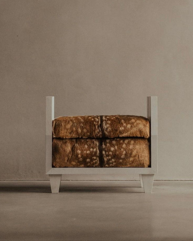The Sycamore Bench - Parchment Seat by Bicci de' Medici, featuring a minimalist design with a textured brown cushion and white wooden legs, is set against a plain beige wall on a smooth floor, evoking the artisanal luxury reminiscent of Jean-Michel Frank for an elegant setting.