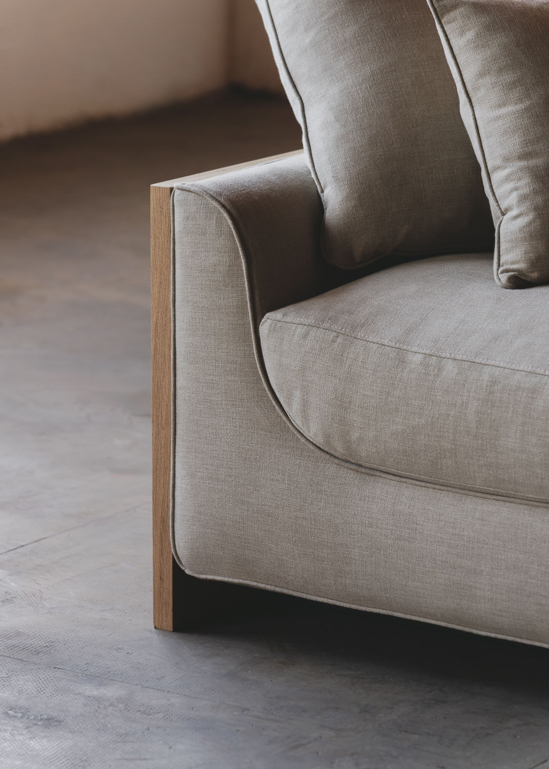 Close-up of ASERIES OF OBSTACLES, SL's Marina Sofa armrest highlights its beige fabric and solid oak frame. Positioned on a concrete floor in a softly lit room, it combines style with comfort through sleek design and removable backrests.