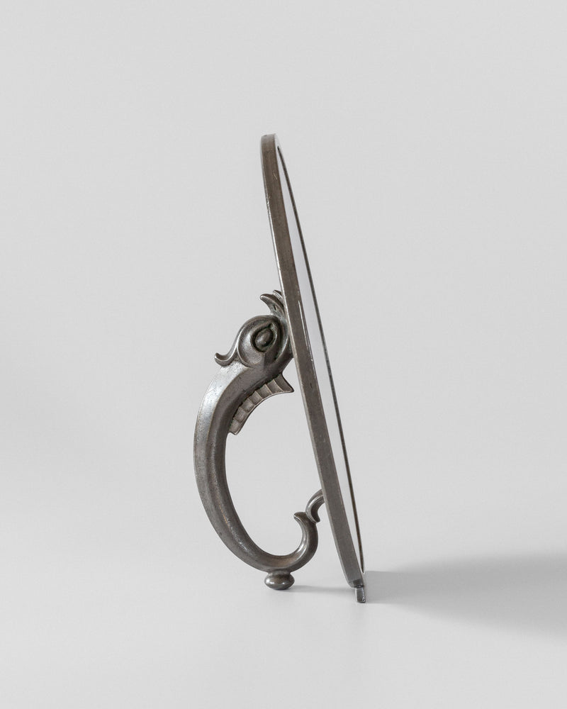 A side view of the Spigel Art Deco Metal Mirror, in vintage condition, highlights its round design set on a decorative metal stand. The ornate stand, evoking the craftsmanship of C.G. Hallberg, is adorned with motifs that resemble a stylized bird or mythical creature, elegantly supporting the mirror against a plain background.
