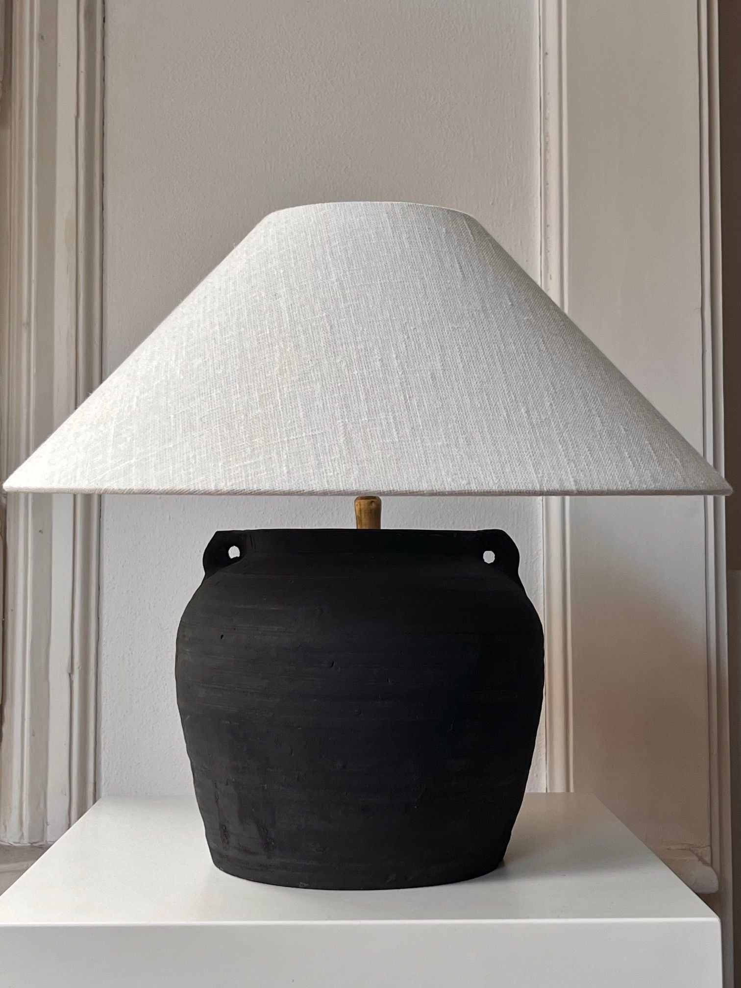 The Médecine Antique Earthenware Lamp, featuring a black ceramic pot and wide, textured white shade, is elegantly displayed on a square white pedestal against a simple white wall backdrop, highlighting its sophisticated black patina.