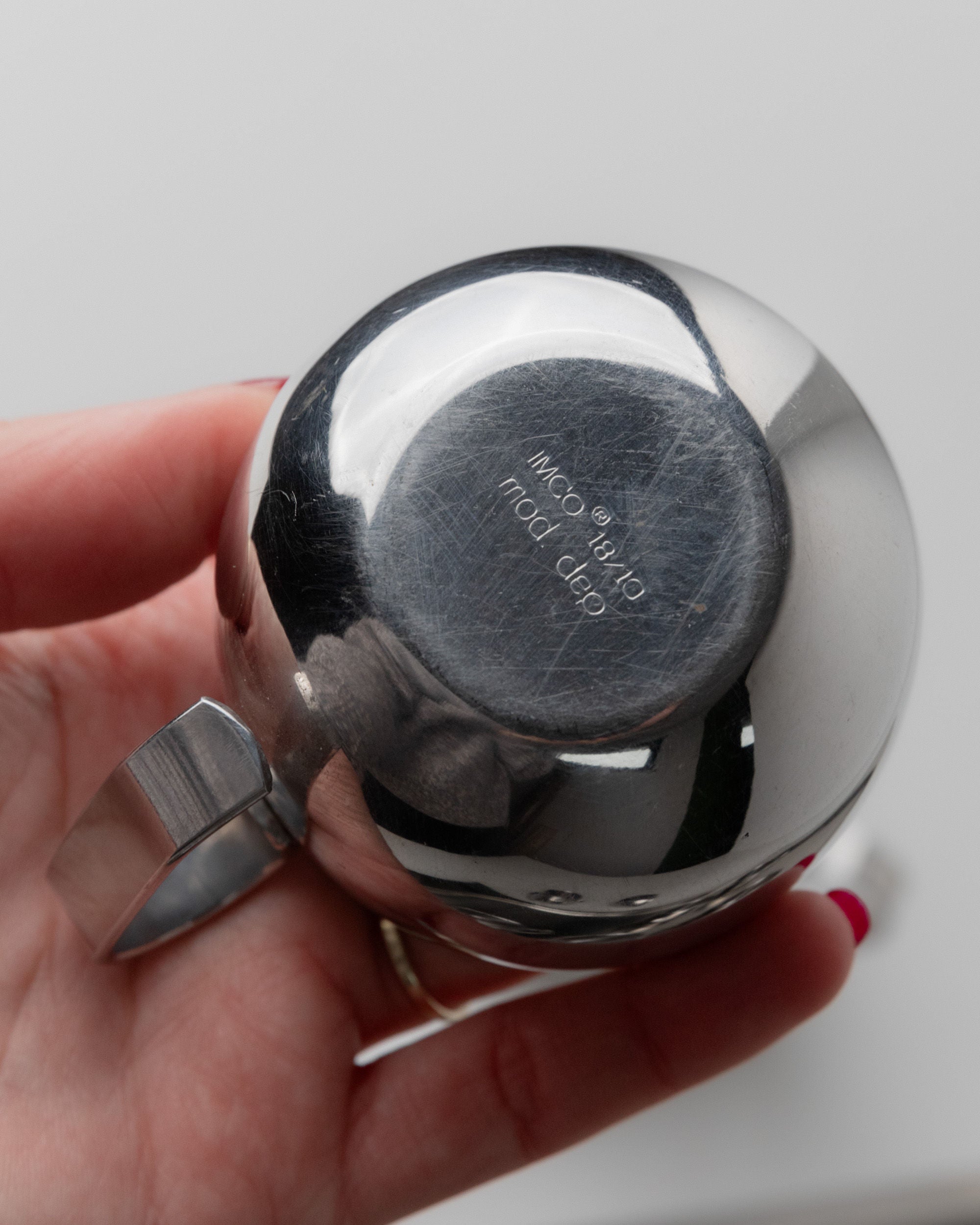 A close-up of someone holding a shiny, round metal object from the 70s Coffee set by IMCO, Bottega Jacobs. It features engraved text on the bottom, a small side handle, and reflects the individual's hand and fingers.