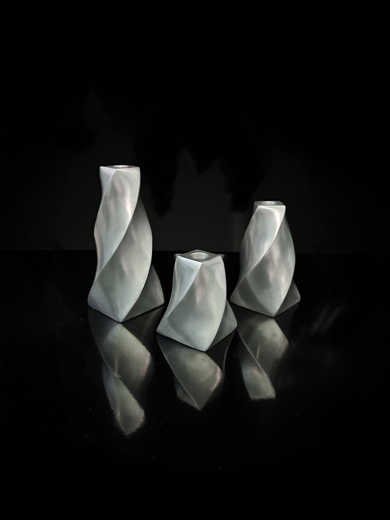 Displayed against a black background are three angular Dodo Vintage candle holders, crafted from aluminium with twisted swirl designs and varying heights. Their metallic surfaces cast delicate reflections on the surface below, evoking an elegant aesthetic.
