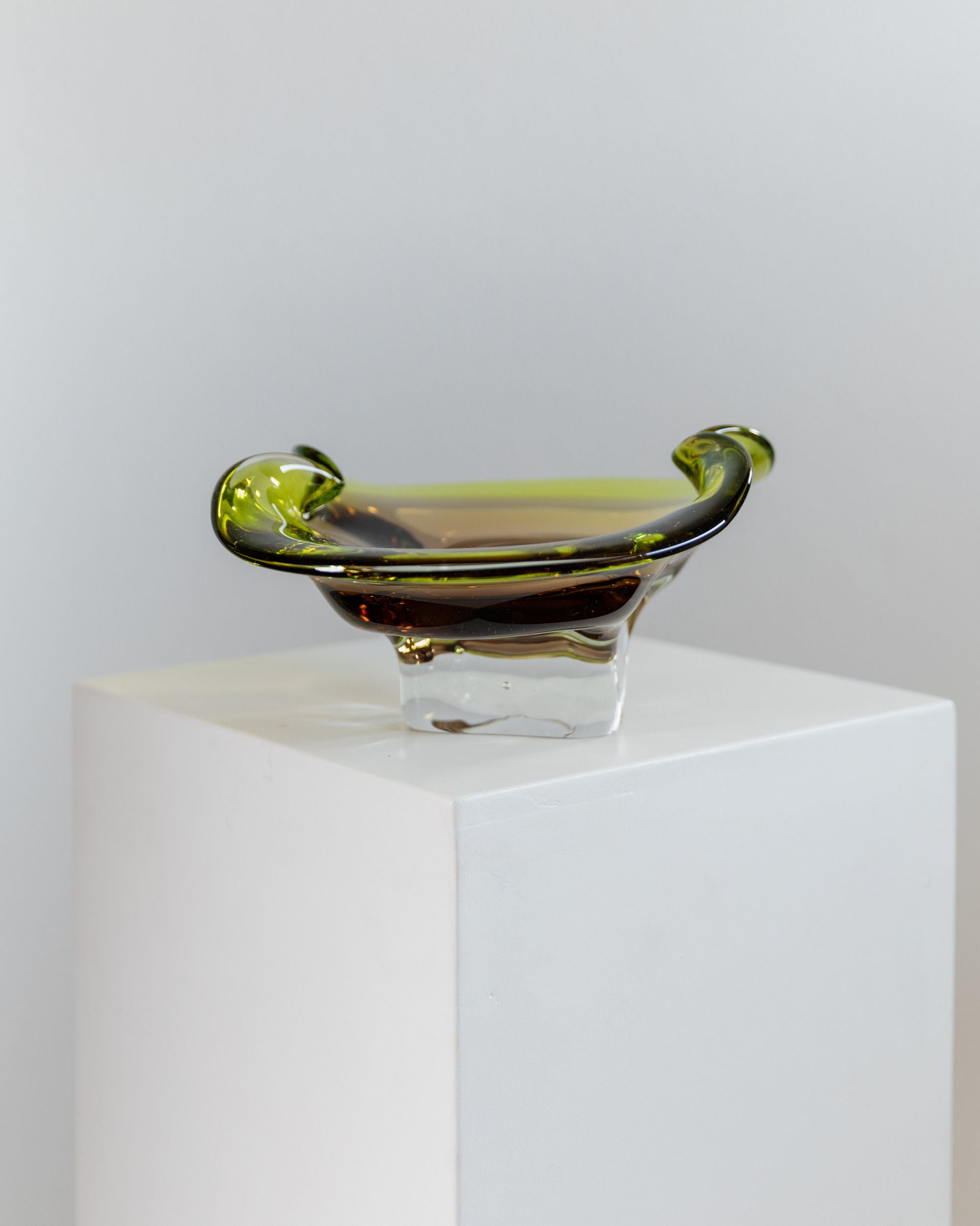 A vintage 1970s Bottega Jacobs Murano Glass Bowl, translucent green with a wavy rim, elegantly graces a white pedestal against a neutral background, highlighting exquisite Italian craftsmanship.