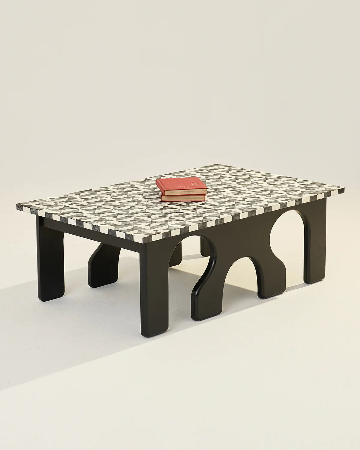 The LMNOH Leo Coffee Table features a modern rectangular design with a checkered black-and-white top and sleek, arch-shaped legs. A red book introduces a splash of color against the neutral, minimalistic background, highlighting the table's refined geometric style.