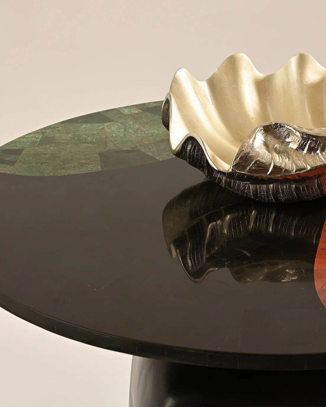 A glossy black LMNOH Jasper Coffee Table is adorned with a decorative metal shell-shaped bowl. Its shiny gold interior and silver exterior complement the semi-precious stones, casting reflections on the table's surface against a plain beige backdrop, enhancing this elegant scene.