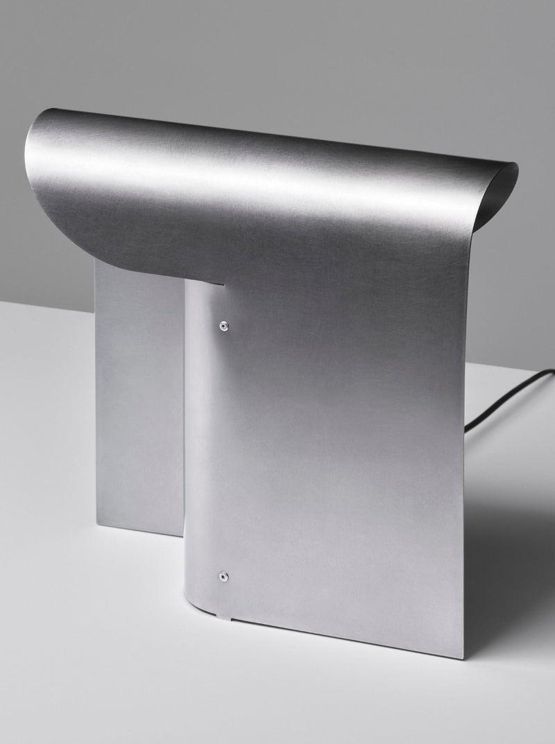 A sleek, minimalist table lamp with a smooth, stainless steel finish. The Arc Table Lamp by Paul Coenen features a curved top and a rectangular base, designed in a contemporary style. The E27 socket ensures versatile bulb compatibility. The lamp is placed on a plain white surface against a neutral gray background.