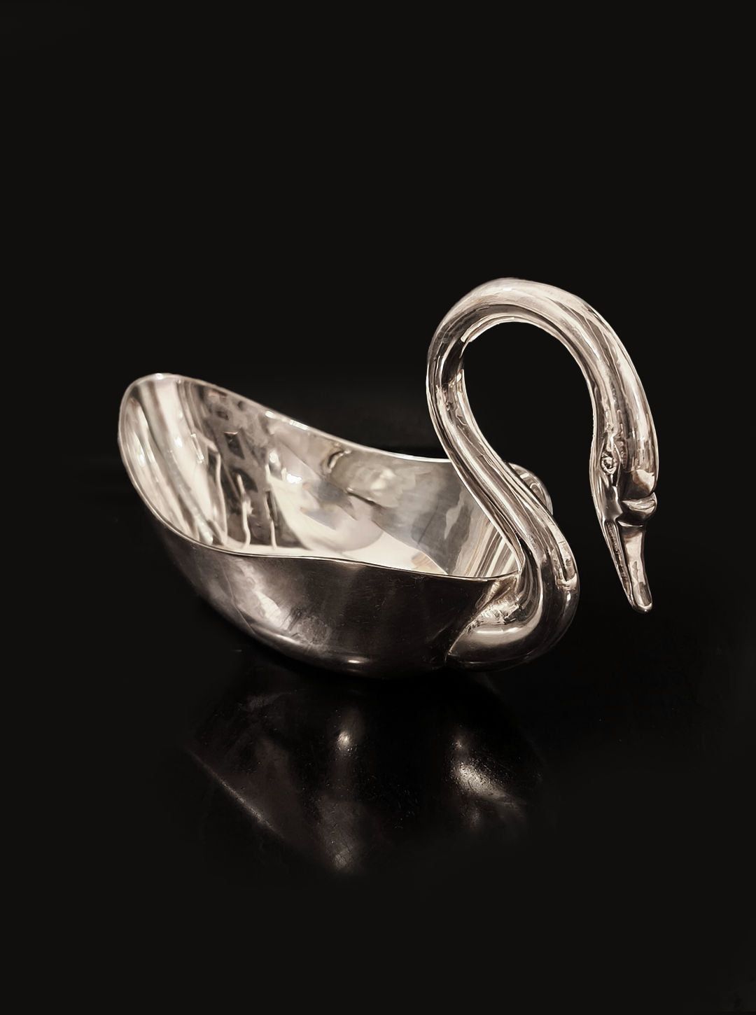 The Swan Silver Saucer by Les Objoies is an elegant decorative piece featuring a graceful, swan-shaped design with a curved neck and head. It rests on a glossy black surface, with its reflective silver finish beautifully highlighting its elegant curves and shimmering appearance.