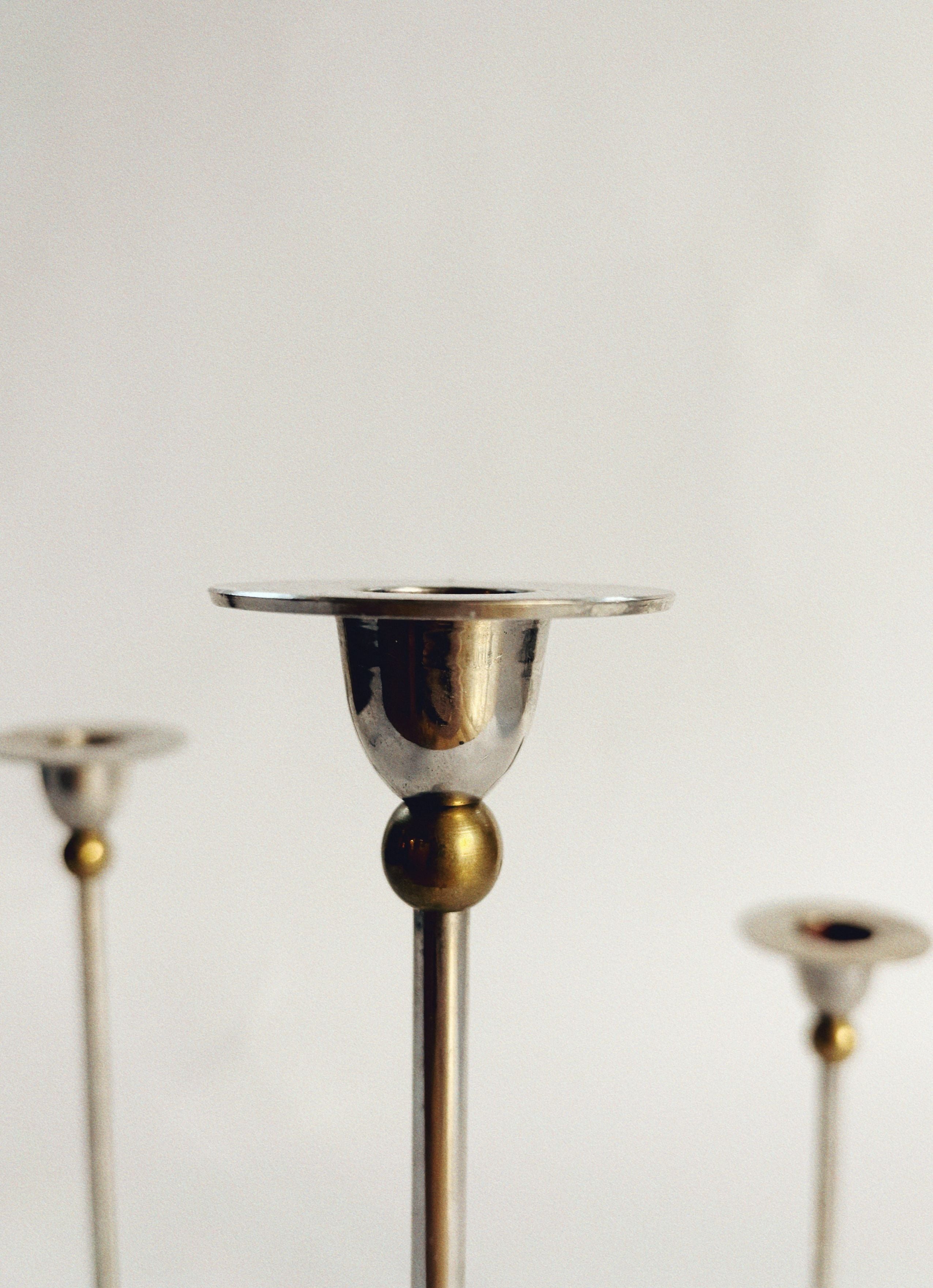 A close-up view of the Set of 3 Postmodern Candle Holders by Huerto showcases their minimalist design, featuring reflective silver cups and brass accents on sleek thin stems, all set against a plain white background.