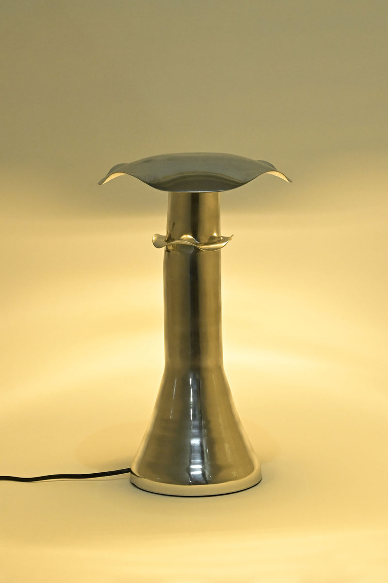 The LMNOH Funghi Table Lamp features a metallic, minimalist design with a flared base, curved top, and left-side black power cord. It emits ambient light in a warm yellow glow.