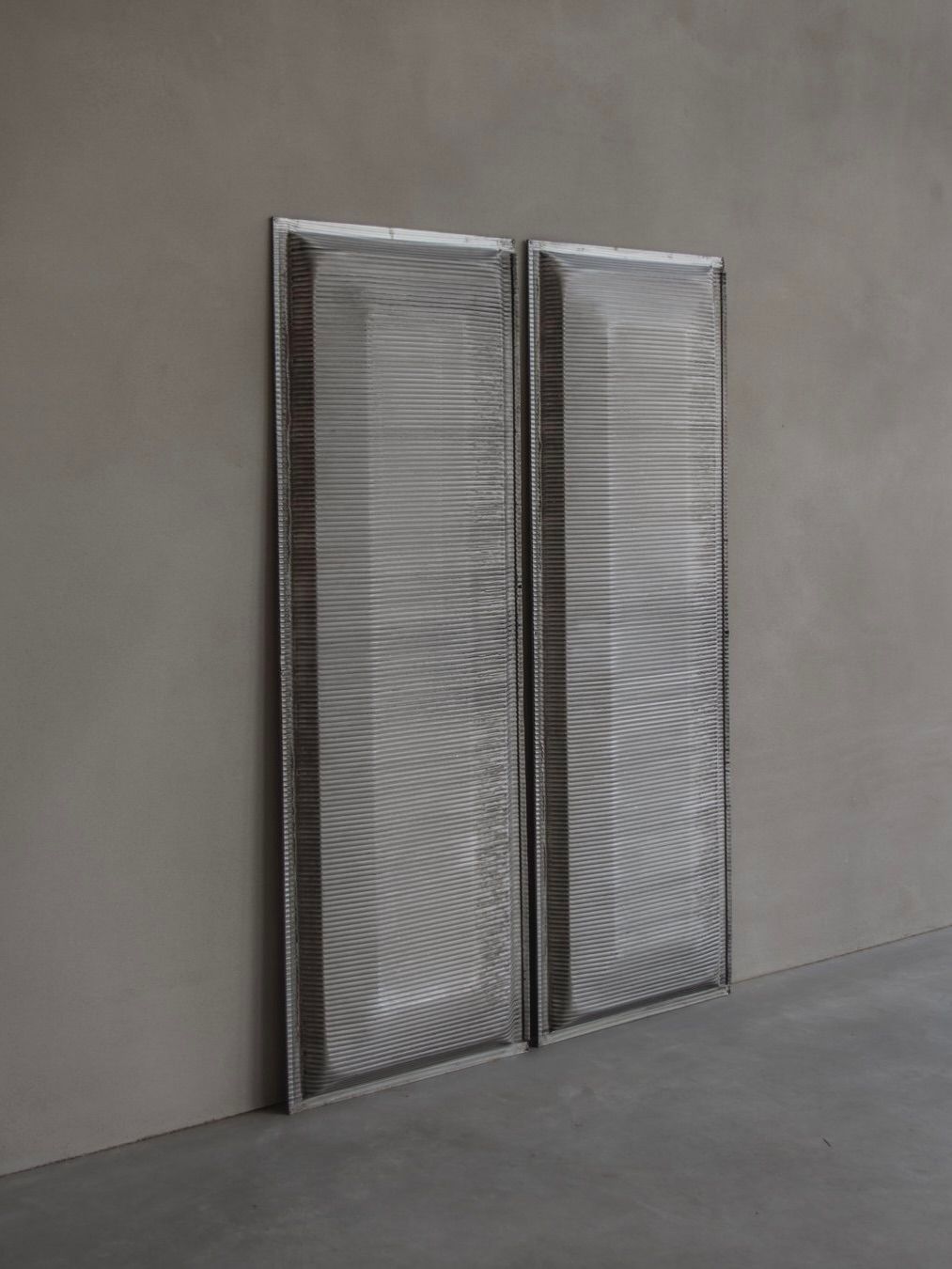 Two tall Jean Prouvé - Façade Panel 70s by Bicci de' Medici, with horizontal ridges, lean against a plain gray wall on a smooth floor. Their equal size and design echo the modern, minimalist style of embossed aluminum grilles.