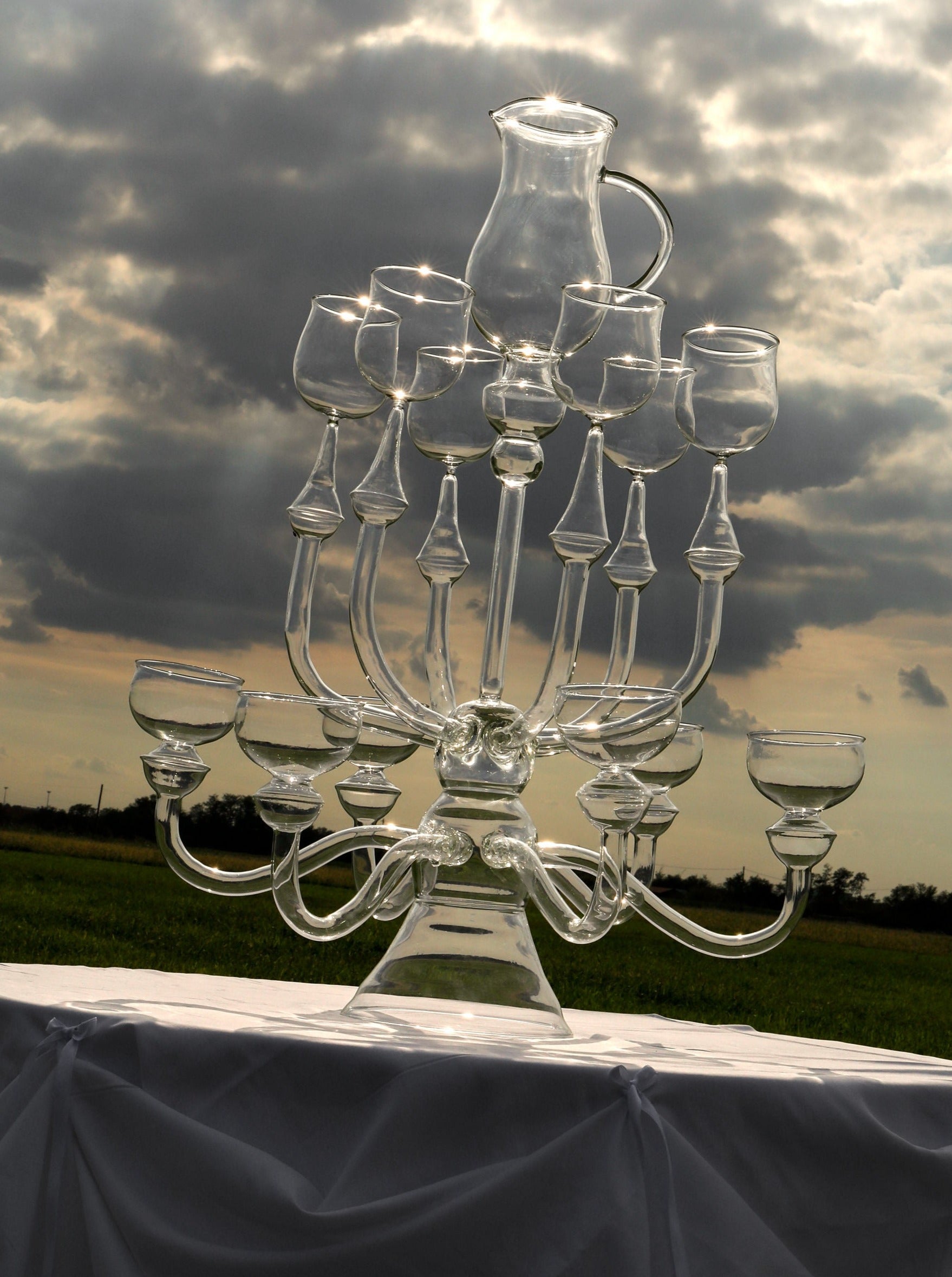 A captivating glass sculpture by Ylaria Pavone named "Dinner’s Ready," featuring a clear candelabrum with 12 arms, each adorned with an empty wine glass and topped with a pitcher, is showcased against a cloudy sky backdrop. This stunning piece is set on a white tablecloth in an open field, providing a unique dining experience with its elegant glass dinnerware.