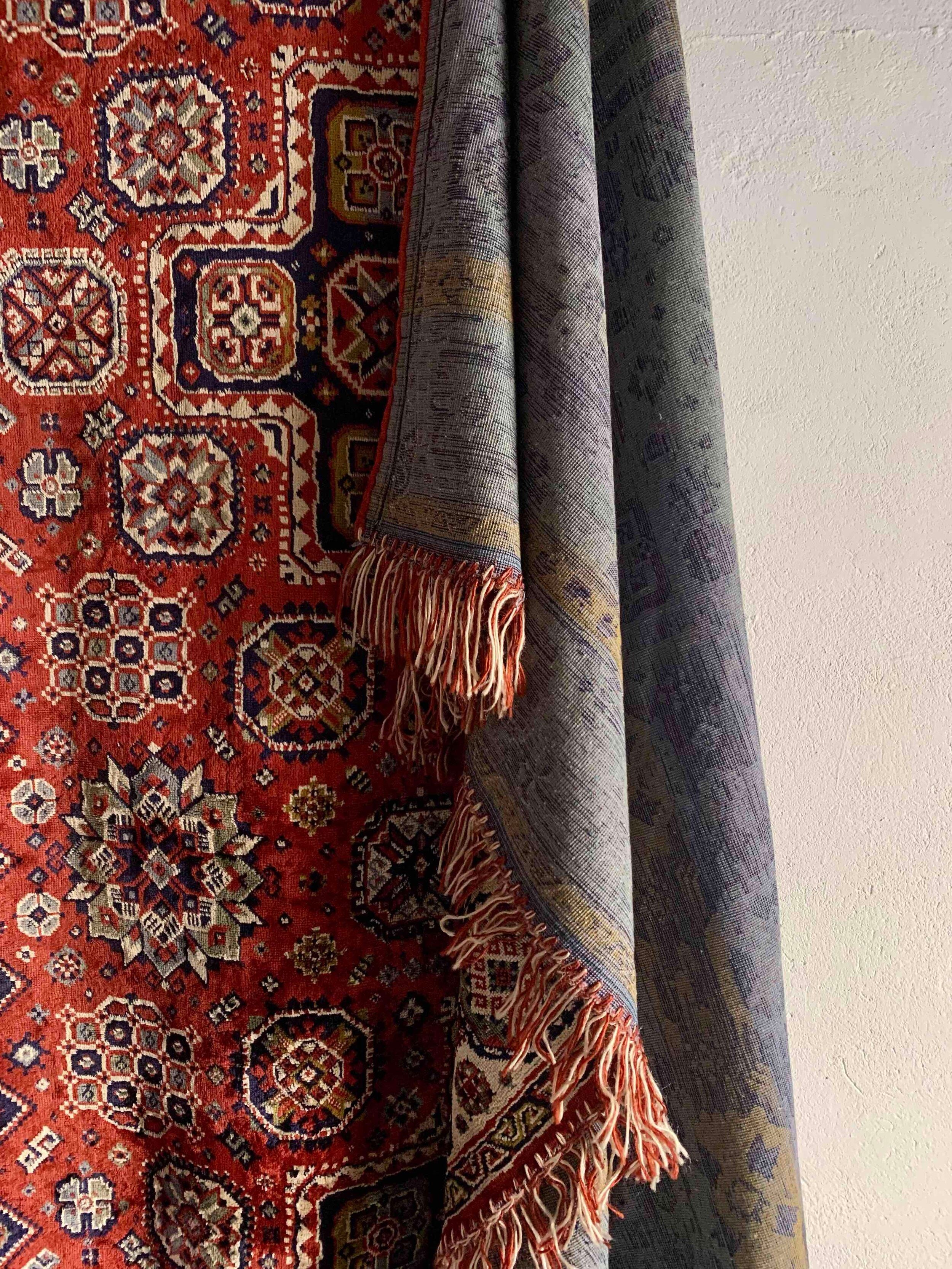 A Veter Vintage Silk Oriental Duvet Cover - Rug with intricate geometric patterns hangs on the wall, while another fringed silk-wool piece in muted tones drapes over it—both showcasing exquisite woven designs.