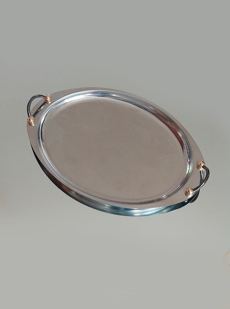 The Medium Oval Steel Tray by Les Objoies is an elegant modern piece featuring sleek, reflective surfaces and a minimalist design. It includes two black handles on either end, each accented with a small golden ball. Its muted gray background enhances its contemporary appeal.