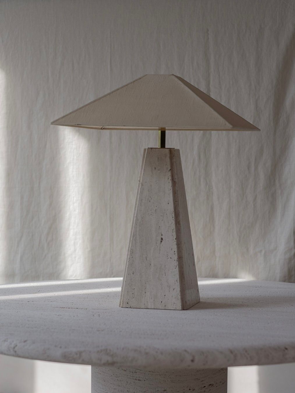 On a round, light-textured table against a neutral backdrop, a minimalist triangular table lamp with a geometric base and beige cone-shaped shade evokes serenity. Its elegant design and modern aesthetic bring to mind the Camille Breesch Travertine Table Lamp by Bicci de' Medici.