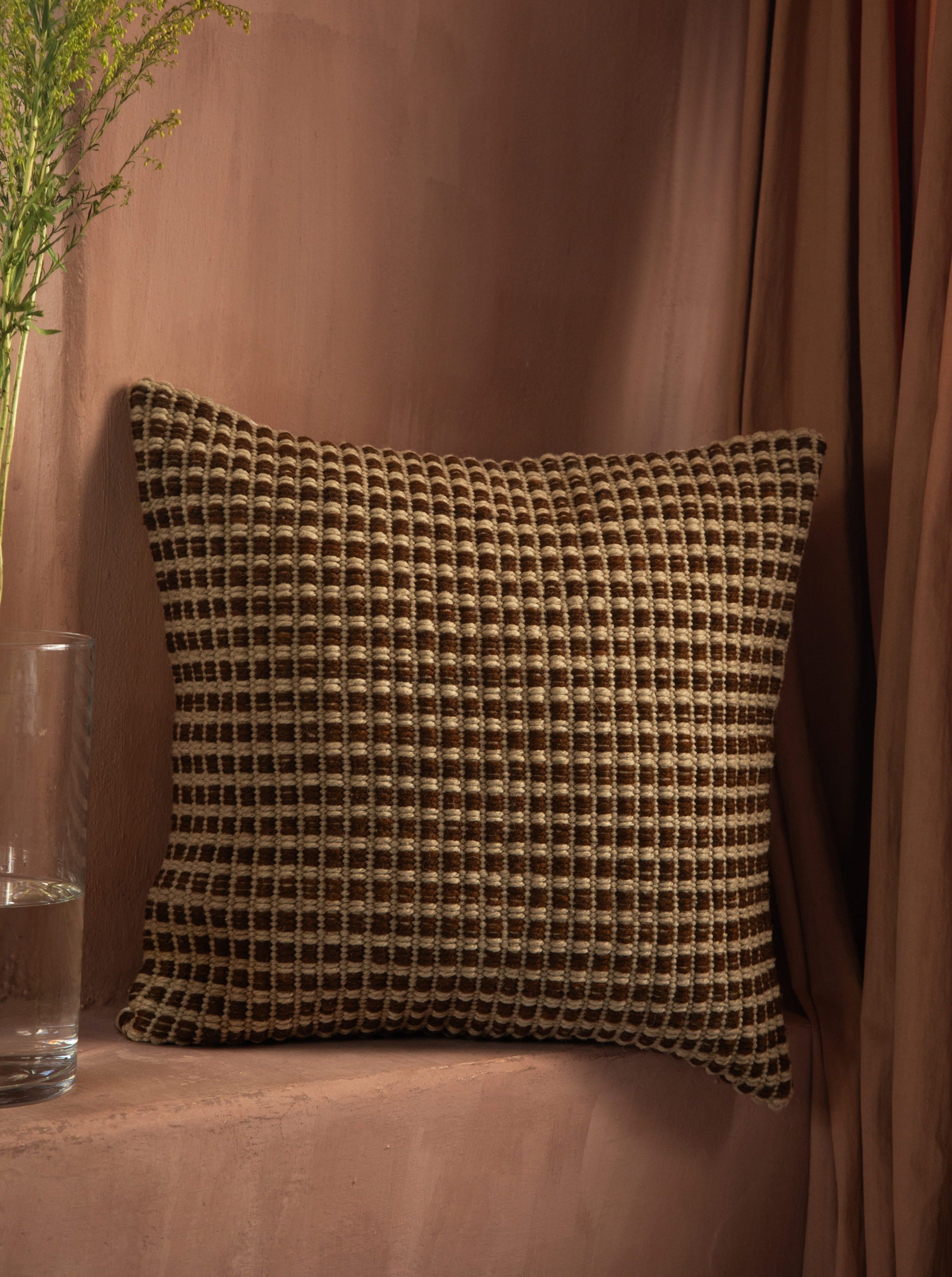 A Identidad Argentina Cima Pillow, crafted from hand-spun sheep wool, against a pink textured wall, with a sheer curtain and a glass of water beside it.