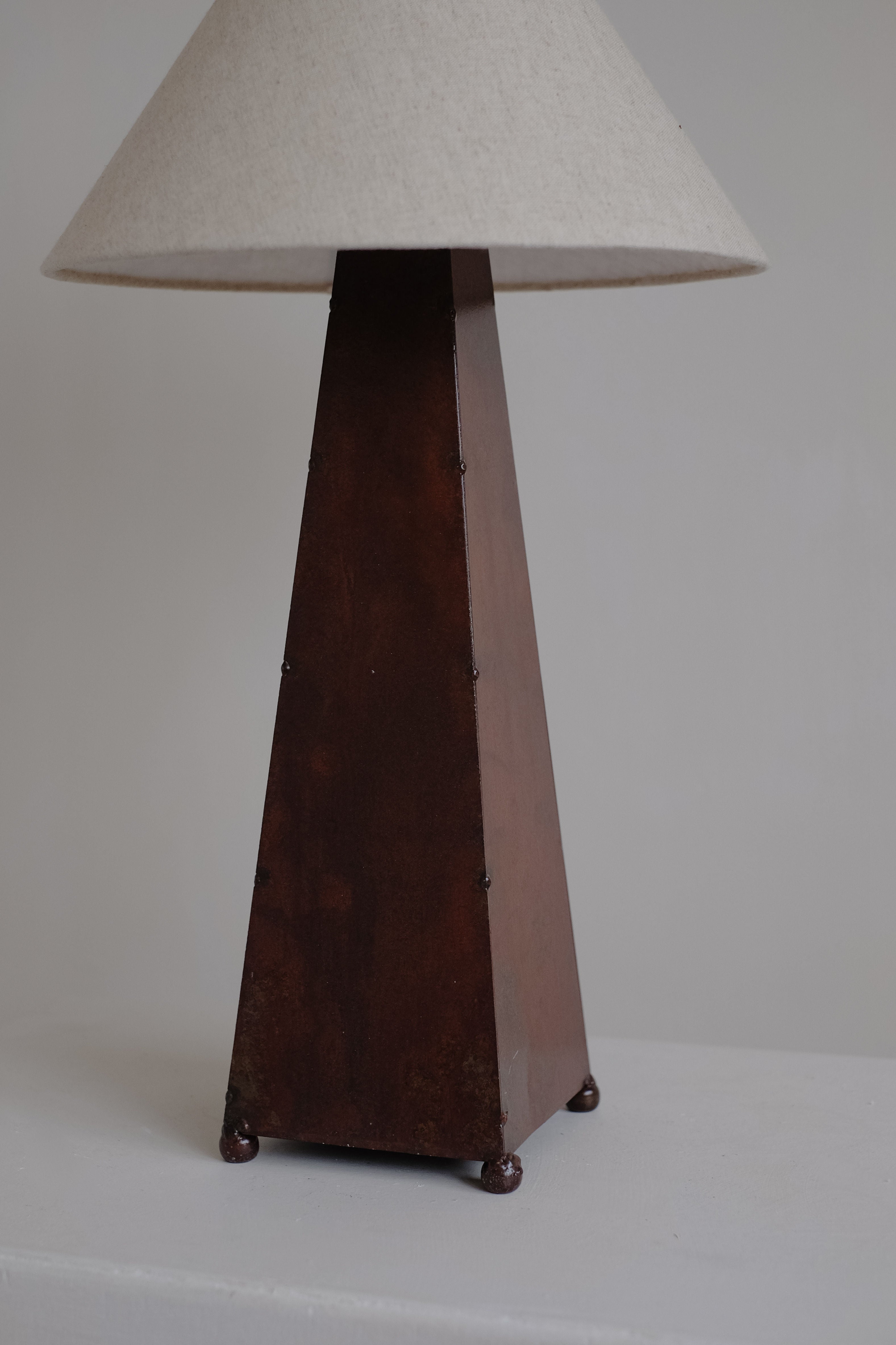 The Obelisk Table Lamp by septembre studios boasts a dark pyramid base with a rust-copper patina, complemented by a textured beige shade. It features a dimmable switch for customizable lighting, making it an elegant addition to any neutral setting.