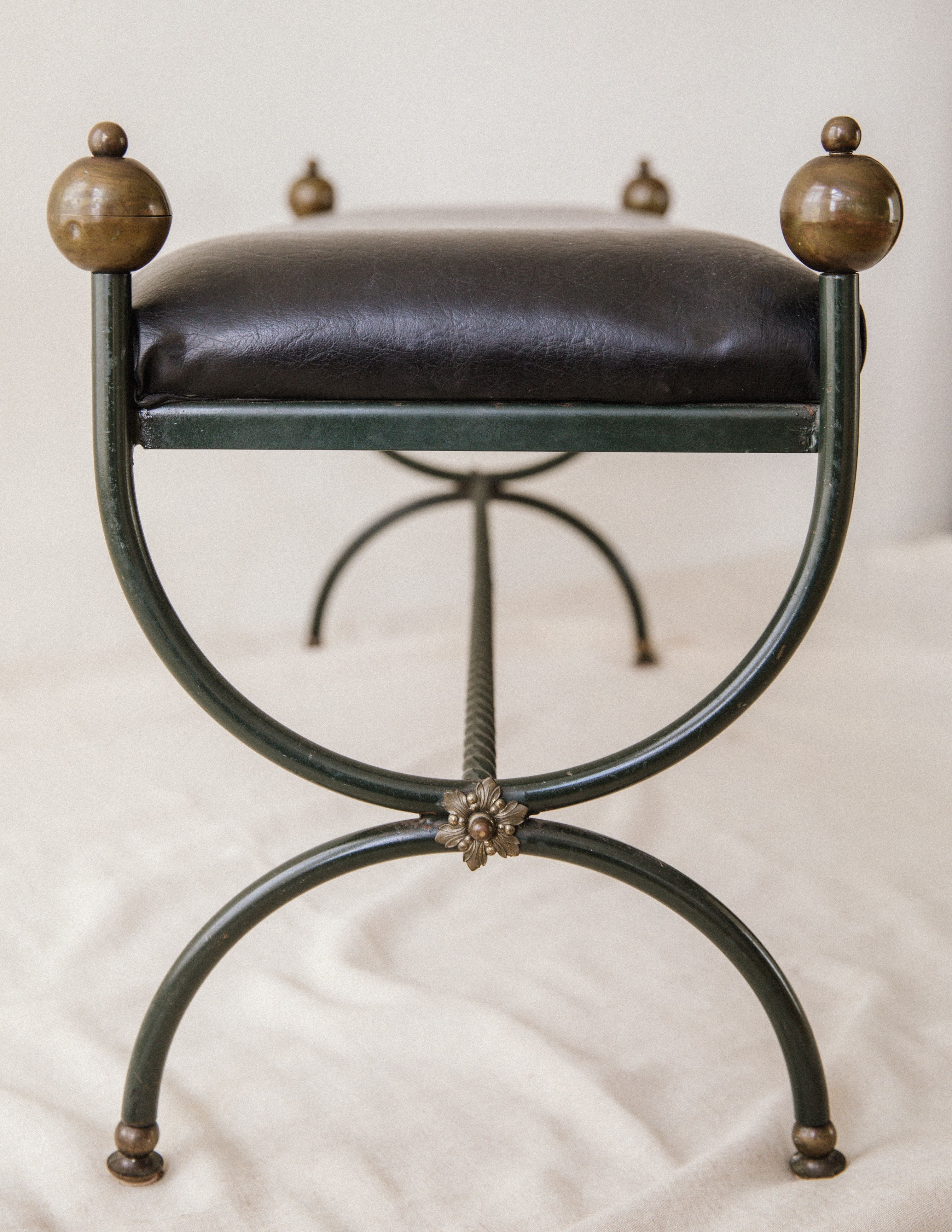 The Arcā Collections Mid-Century Wrought Iron Bench features a black cushioned seat, metal frame with decorative brass knob finials, curved legs, and a floral accent at the joint, shown on a light fabric surface.