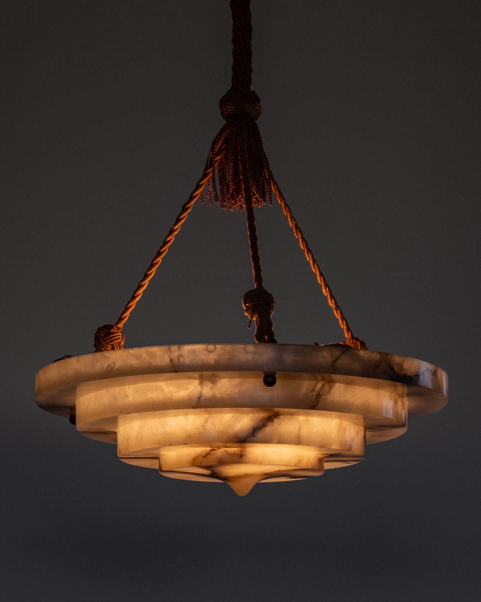 The Spigel Art Deco Alabaster Pendant Chandelier features layered, circular marble-like shades and is suspended by braided ropes against a dark backdrop, reminiscent of Swedish Grace design. It emits a warm glow for a cozy ambiance.