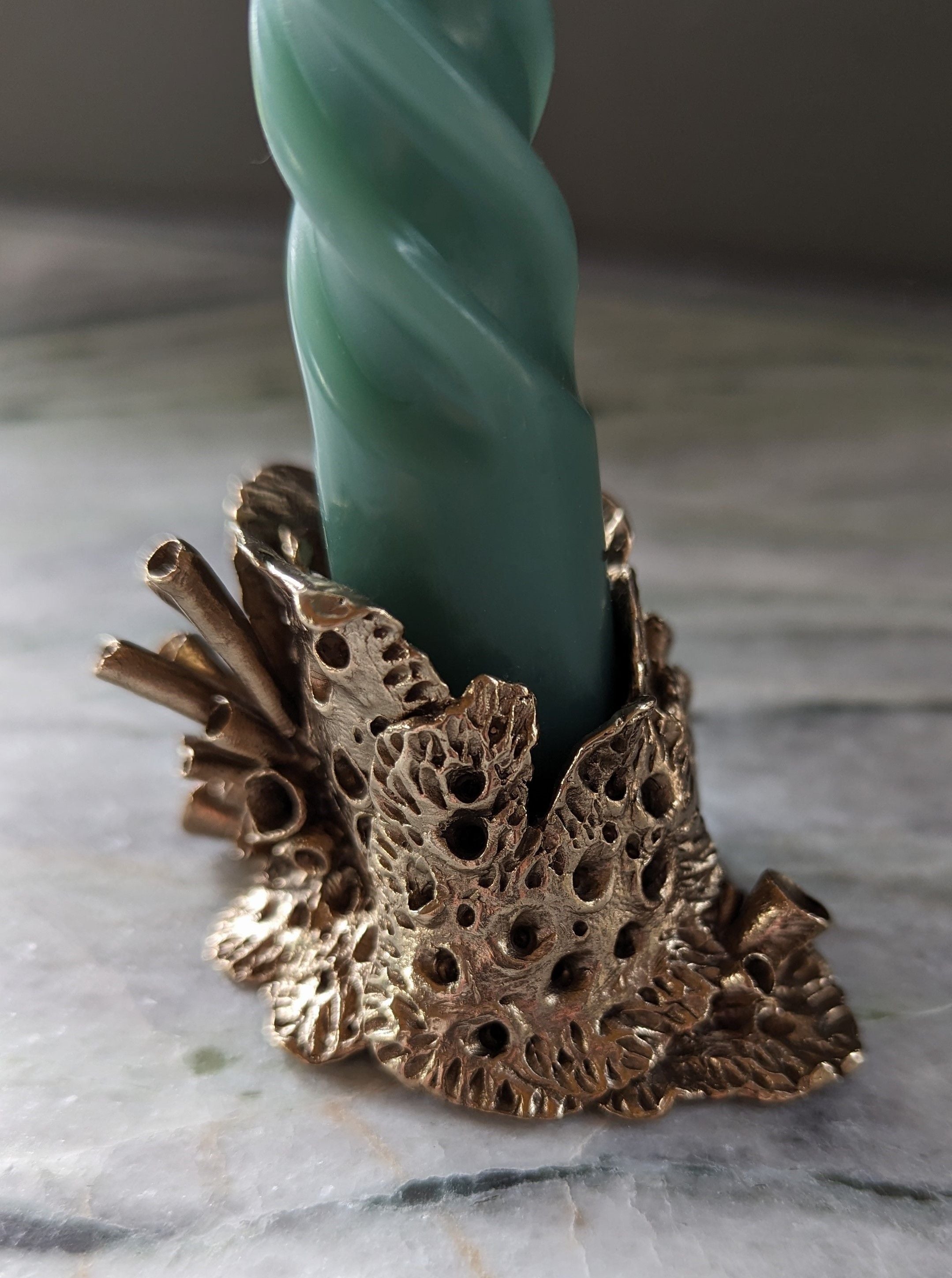 A green, twisted candle standing upright in a Bronze Lichen Candle Holder featuring detailed leaf designs, created using lost wax casting, set on a marble surface by Sophie Symes.