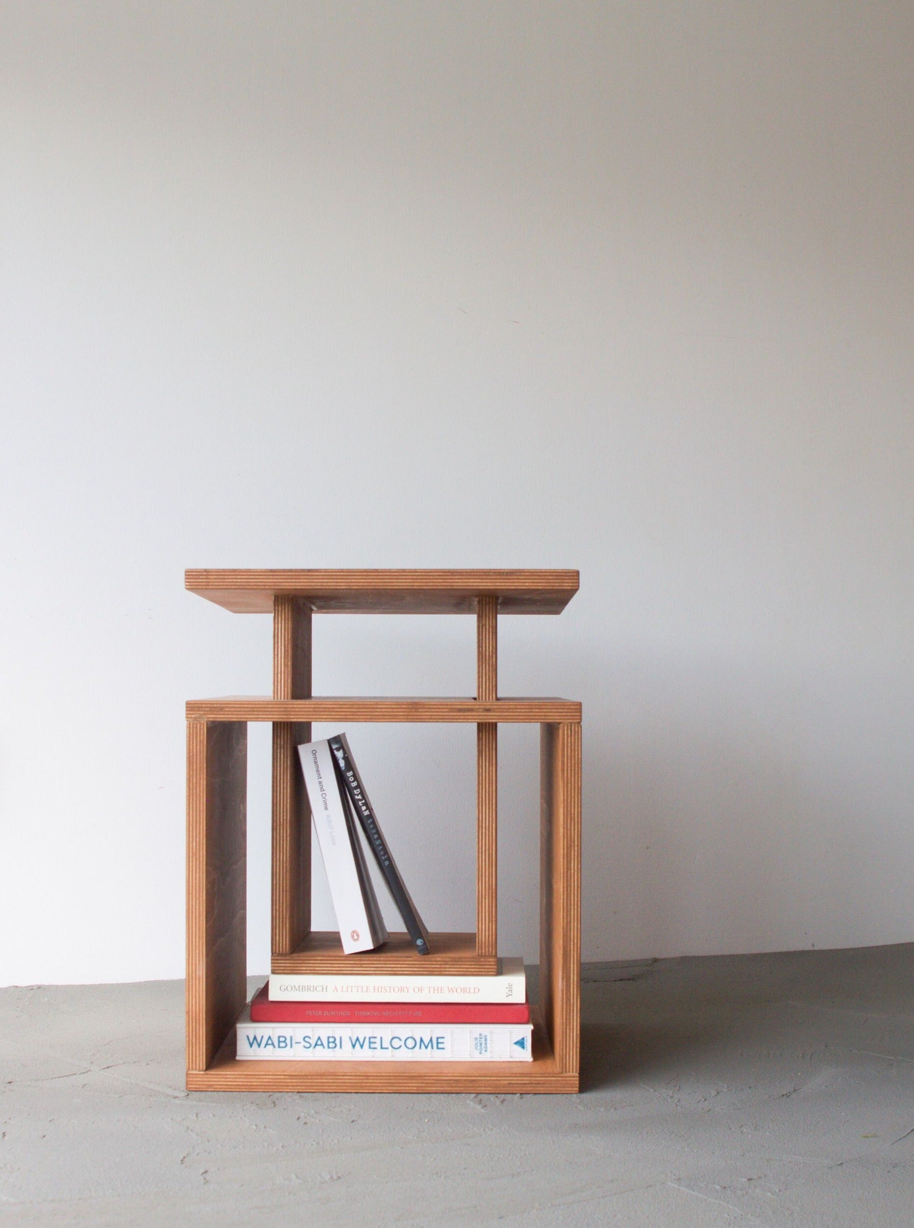 A minimalist Cubo side table with two shelves, containing a couple of books, positioned against a plain white wall on a gray floor by Edoardo Lietti Studio.