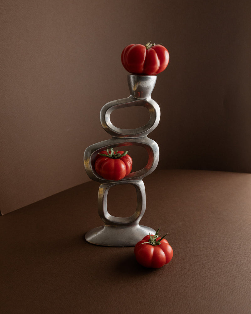 Three red tomatoes are artfully placed on the abstract, geometric Matthew Hilton Candleholder 70s by Bottega Jacobs, with its silver finish reminiscent of mid-century design against a brown background. Two tomatoes rest at different levels on the structure, while one sits elegantly on the surface beside it.